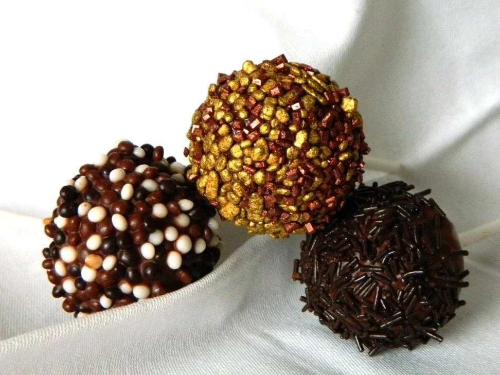 Three truffles decorated with colorful sprinkles, chocolate, and edible gold, resting on white satin fabric