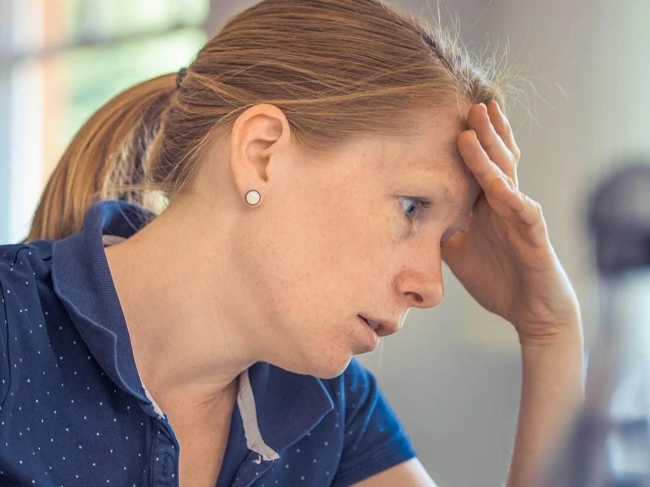 A woman with red hair and freckles holding her forehead, looking concerned or stressed, wearing a blue collared shirt and earrings.