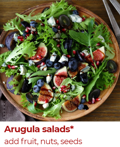 A wooden bowl filled with an arugula salad containing figs, blueberries, black olives, pomegranate seeds, and slices of cheese, on a wooden table with a gray napkin and silverware nearby.