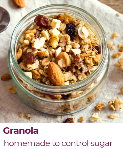 A glass jar filled with homemade granola containing oats, nuts, dried fruits, and seeds, placed on a gray fabric surface.
