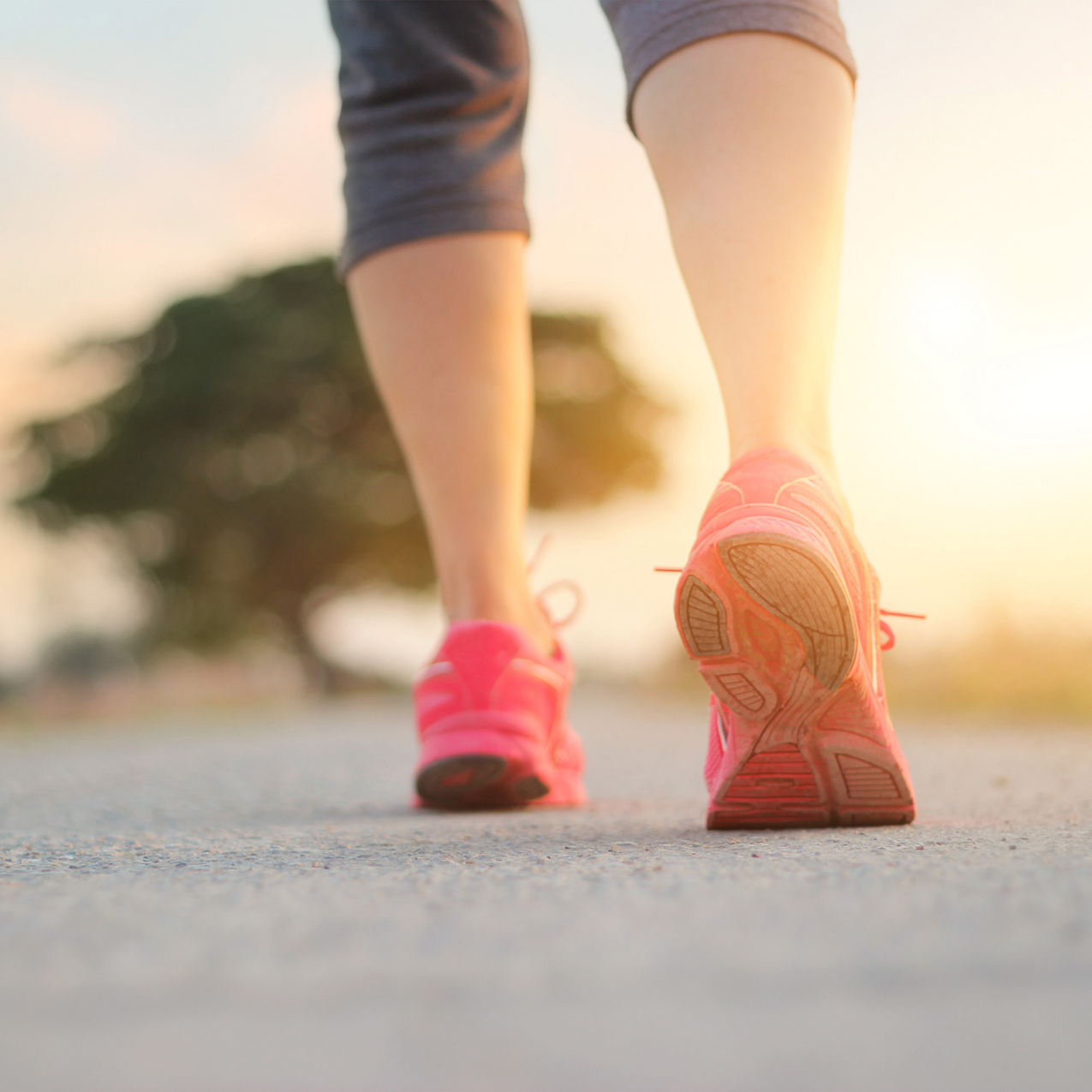 Close-up of a person jogging outdoors on a paved surface during sunrise, wearing pink running shoes and gray athletic shorts.