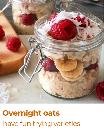 A glass jar filled with overnight oats topped with raspberries, banana slices, shredded coconut, and a drizzle of yogurt or cream. In the background, another jar with similar contents is visible, along with a wooden cutting board with a raspberry on it.