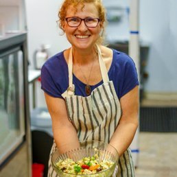 A woman with curly red hair and glasses smiling, wearing a striped apron, holding a bowl of salad in a kitchen.