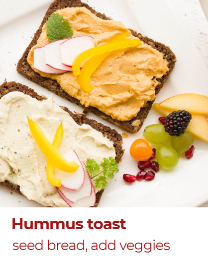 Two slices of seed bread topped with hummus, radish slices, yellow bell pepper slices, and green herbs, garnished with fresh fruit including blackberries, green grapes, and a pear
