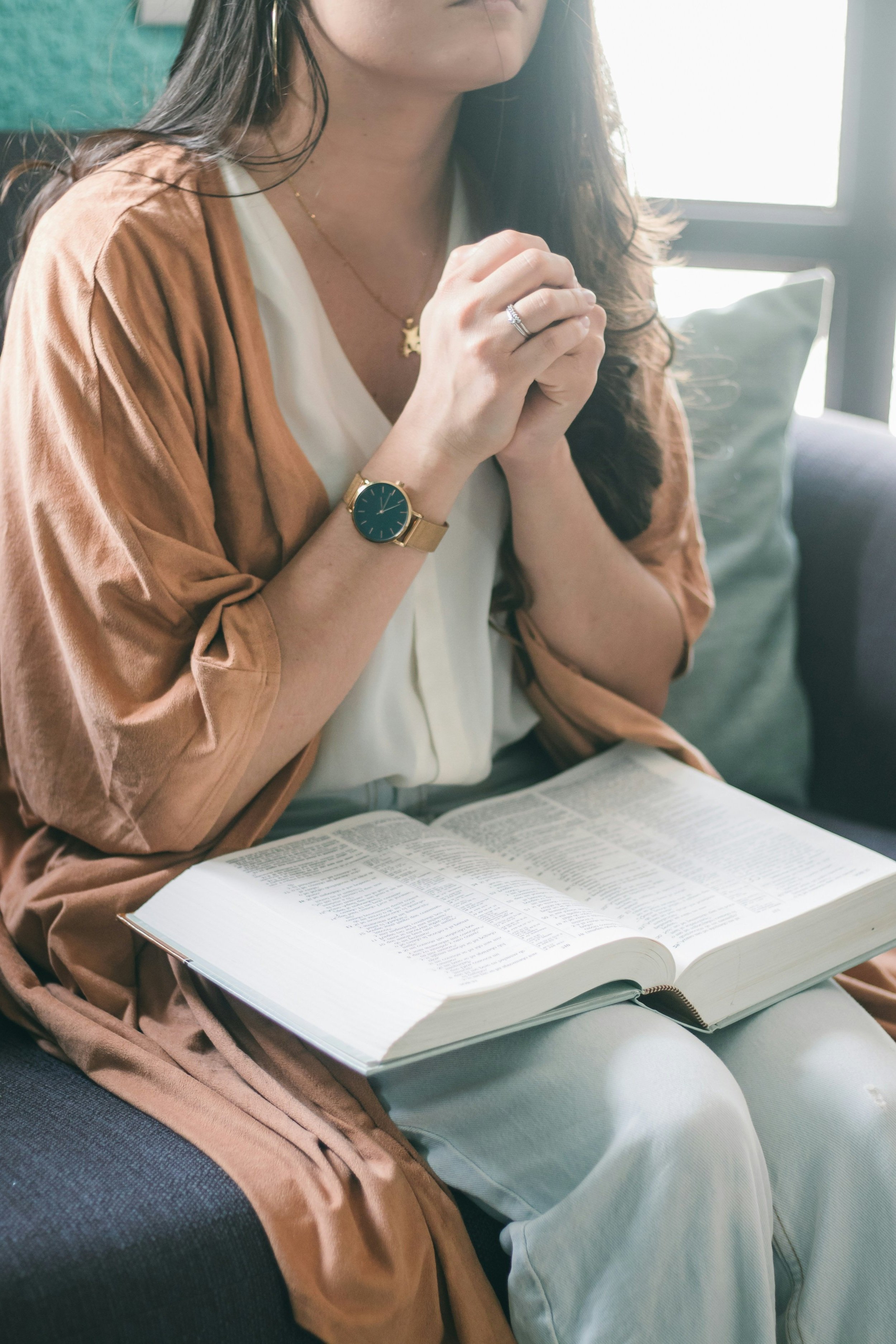 A woman sitting on a dark gray couch, wearing a white blouse and a brown jacket, with a gold watch, a ring, and a gold necklace. She has her hands clasped together in a prayerful or meditative position. An open book is resting on her lap, and a gray cushion is visible behind her. Bright light coming from a window illuminates the scene.