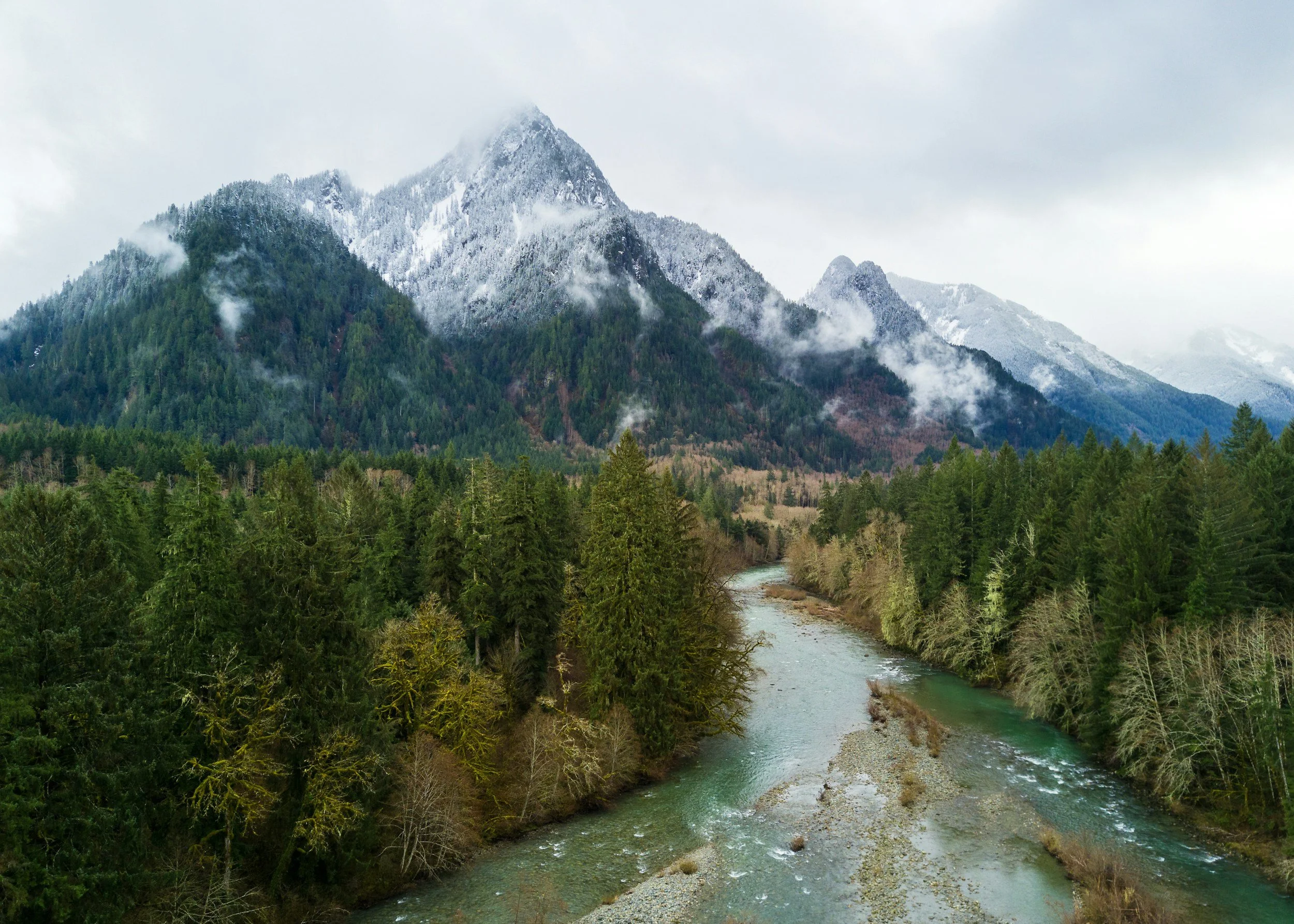 A mountain landscape with snow-capped peaks, a forest of evergreen trees, and a river flowing through the valley.