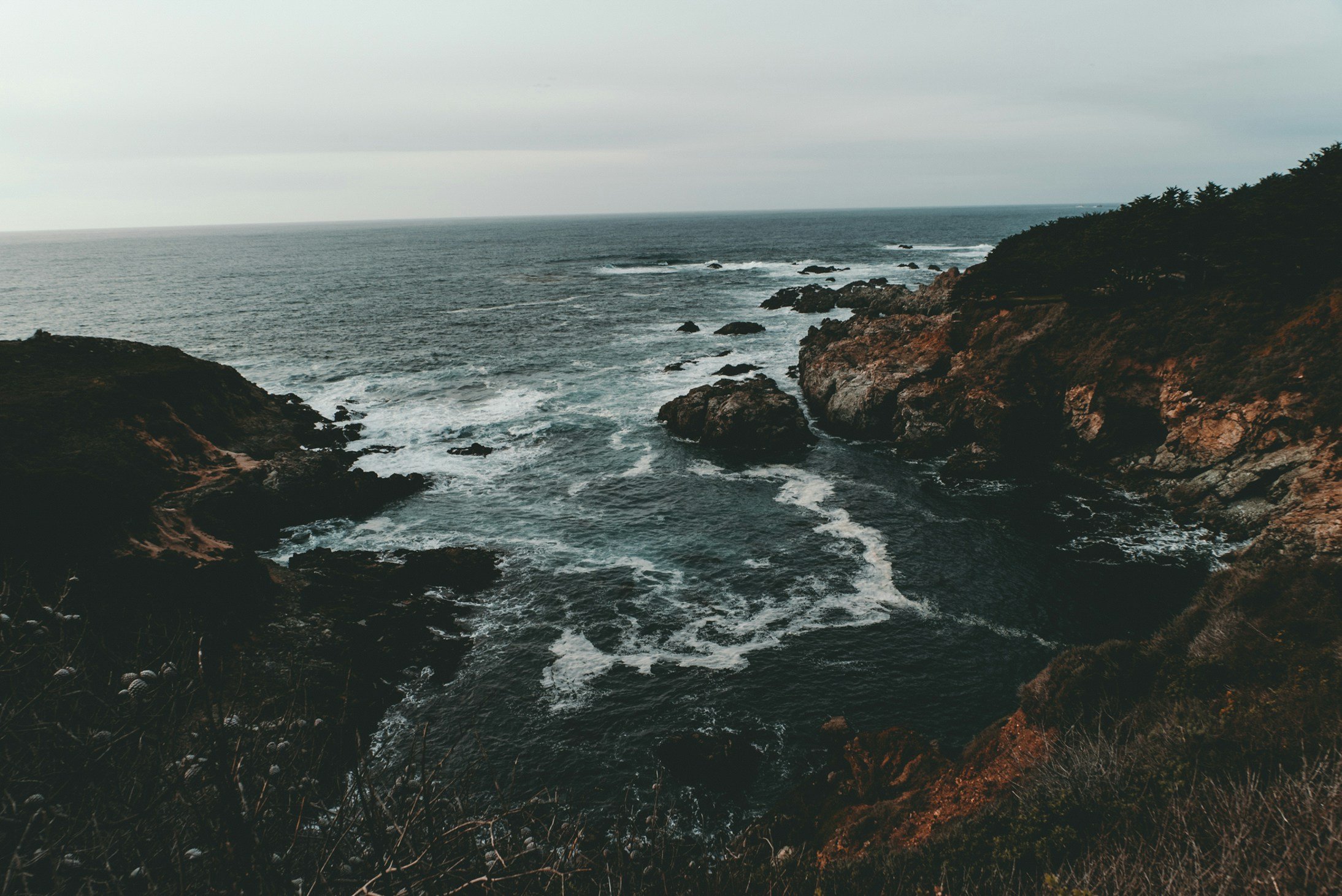 A view of the ocean with waves crashing against rocky cliffs under an overcast sky.