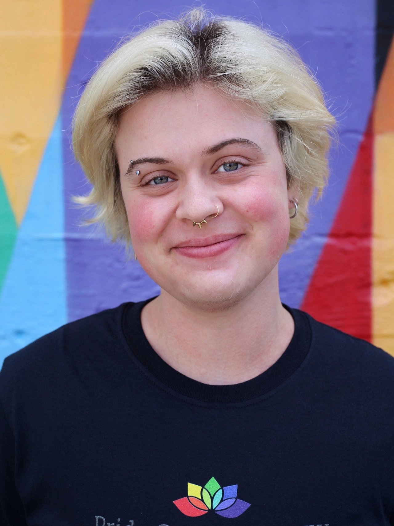 A young person with blonde hair and multiple facial piercings, smiling, standing in front of a colorful, rainbow-like mural.