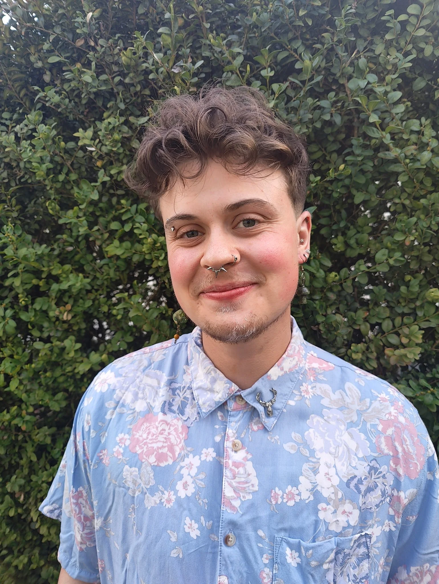 A person with short hair and multiple facial piercings, including a septum and eyebrows, smiling at the camera 