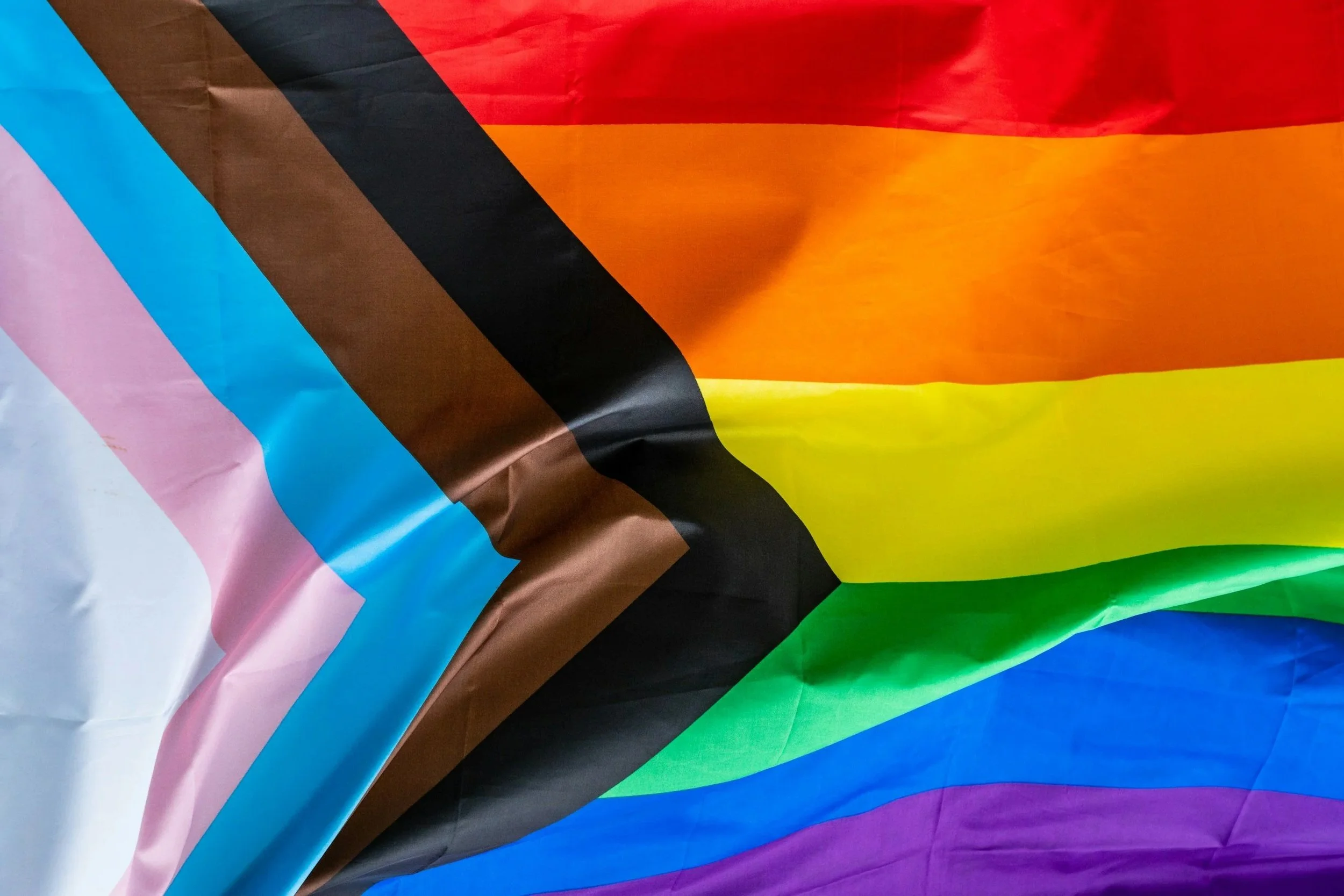 Close-up of a colorful Pride flag featuring pink, blue, brown, black, red, orange, yellow, green, blue, and purple stripes, with some wrinkles and folds.