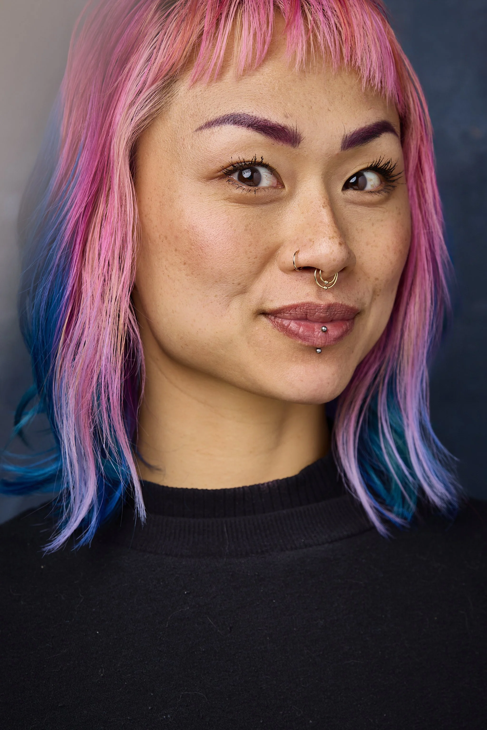 Close-up of a young woman with pink and blue ombre hair, light skin with freckles, and multiple facial piercings, wearing a black top.