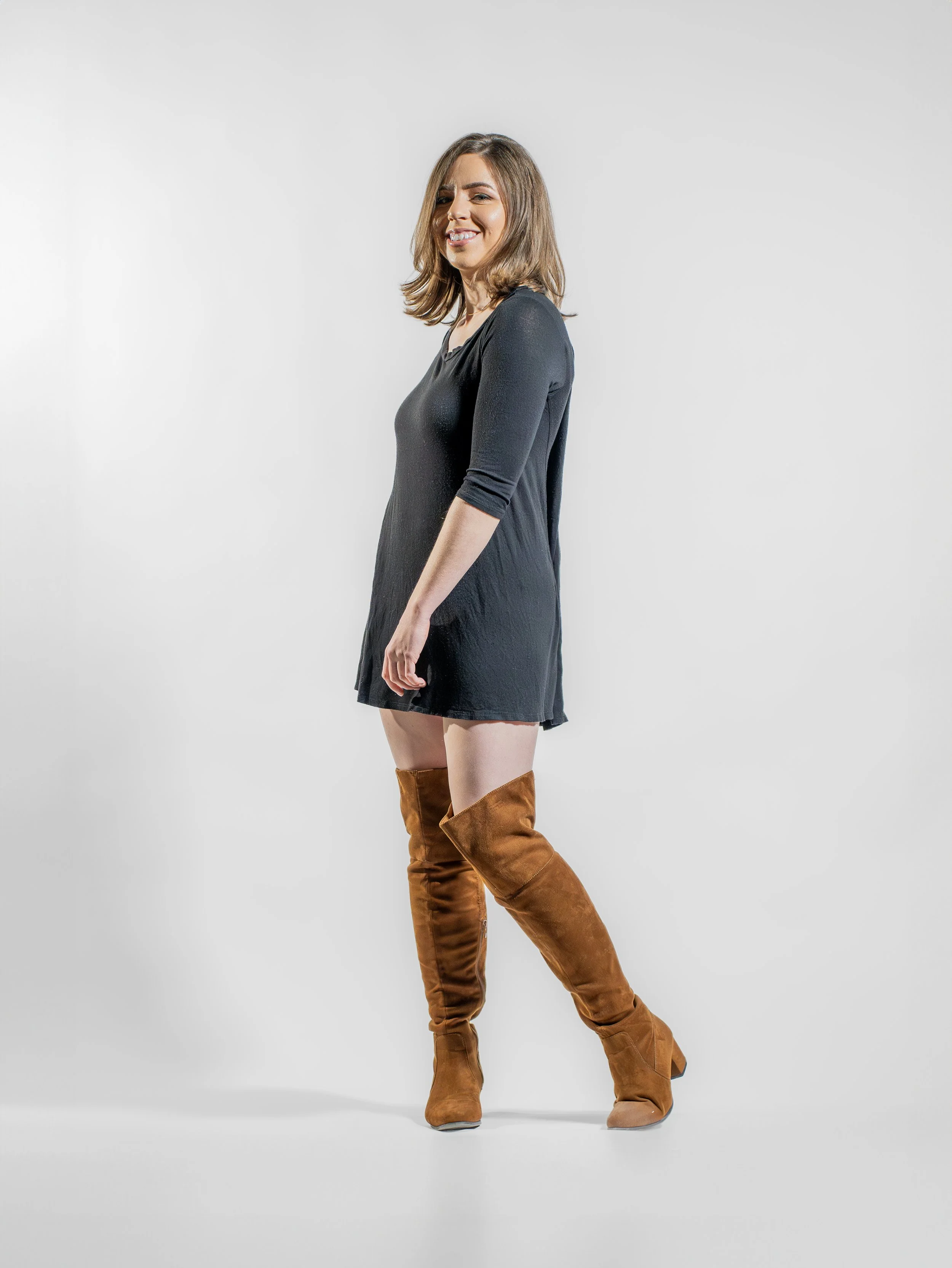 A woman with shoulder-length brown hair standing against a plain white background, wearing a black dress and brown knee-high boots, smiling and looking at the camera.