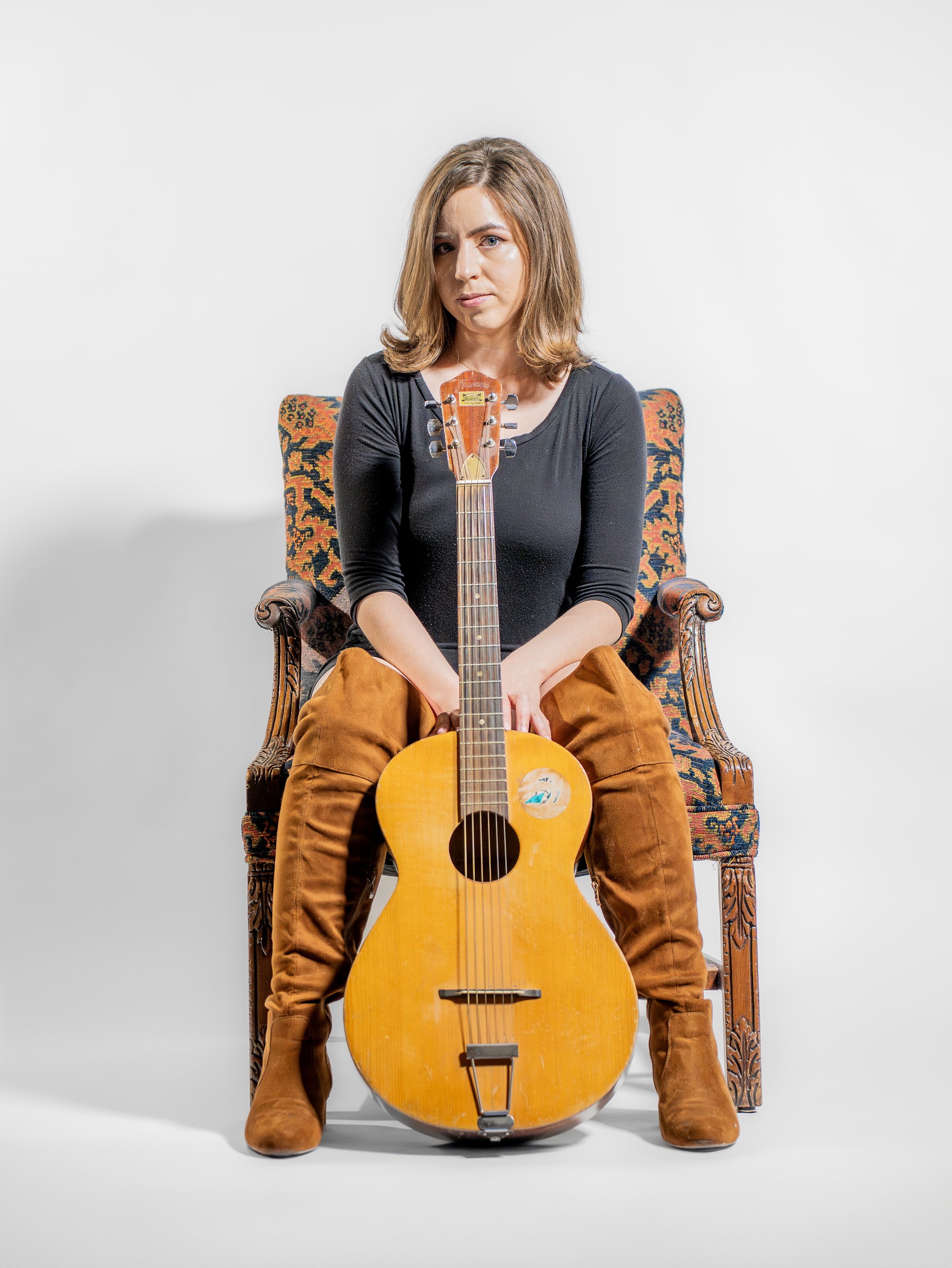 A woman sitting on an ornate wooden chair with a vintage guitar between her legs, against a plain white background.