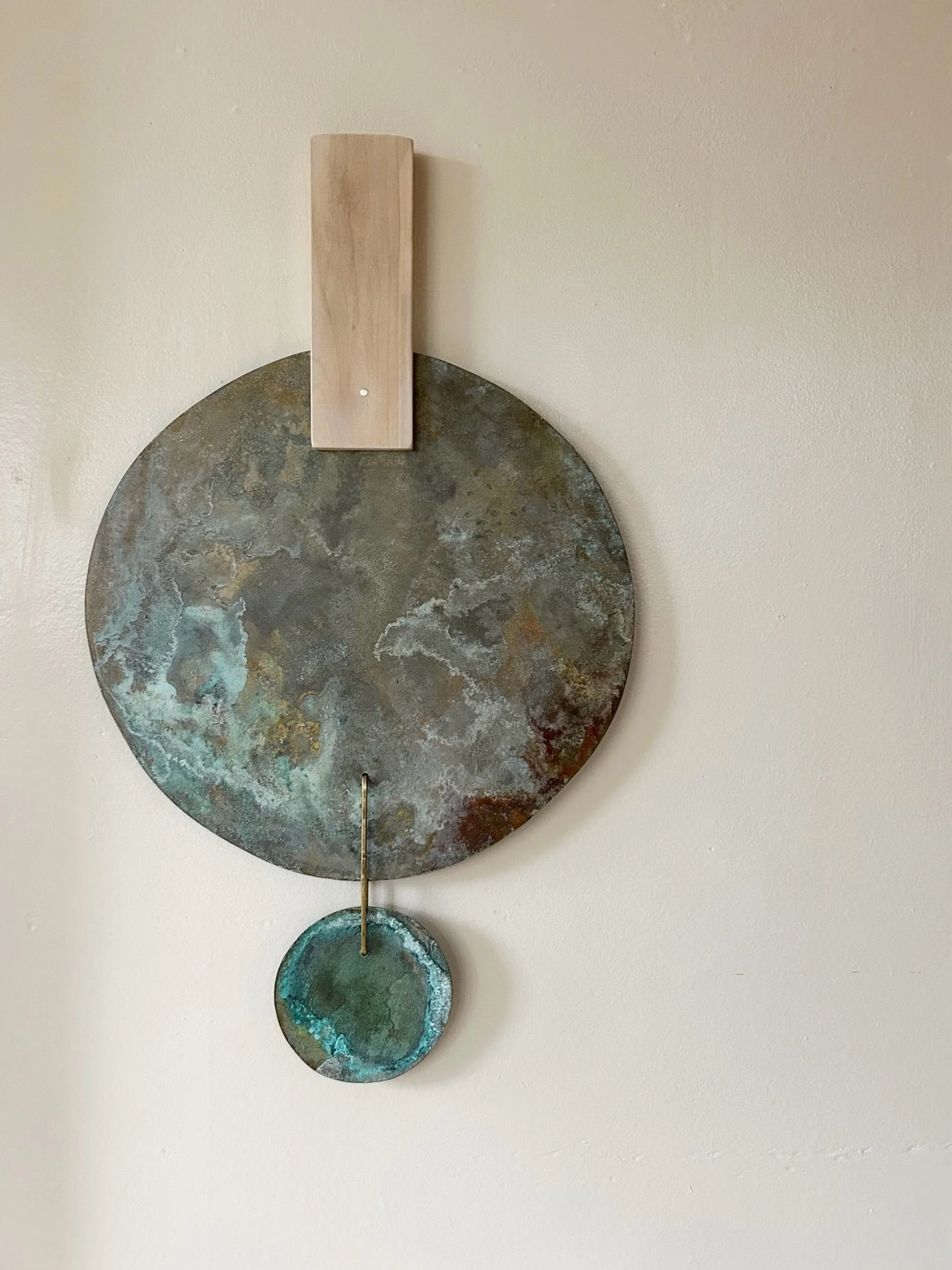 Decorative metal wall art with a wooden hanger, featuring a large round textured metal circle with a smaller round hanging piece, all mounted on an off-white wall.