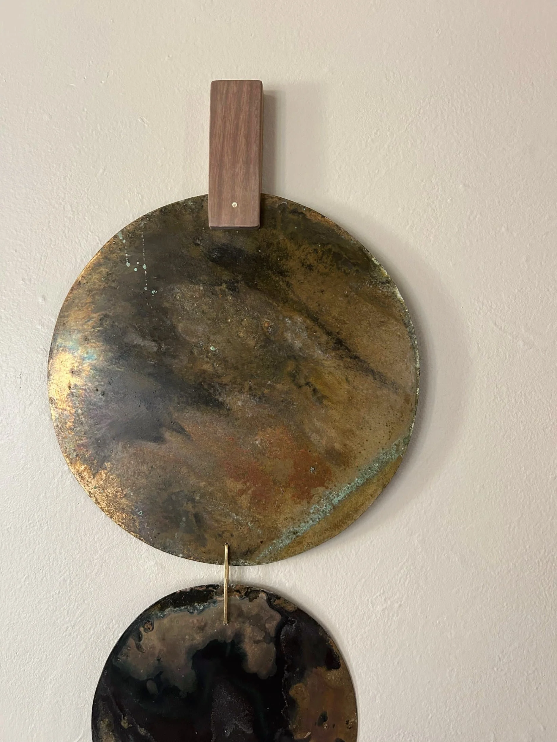 Two metallic plates with oxidized, rusted, and darkened surfaces hanging vertically on a wall, connected by a small hook. The upper plate is oval with a patina of various browns, black, and hints of green. The lower plate is smaller, darker, and more