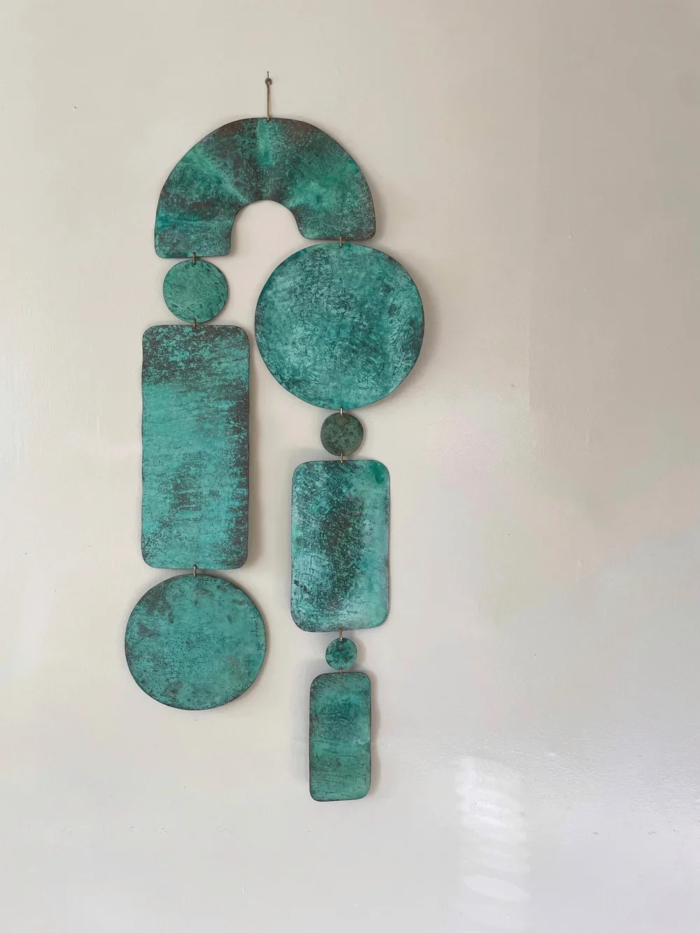 A decorative metal wall art piece with various geometric shapes in a blue-green patina, arranged vertically on a plain white wall.