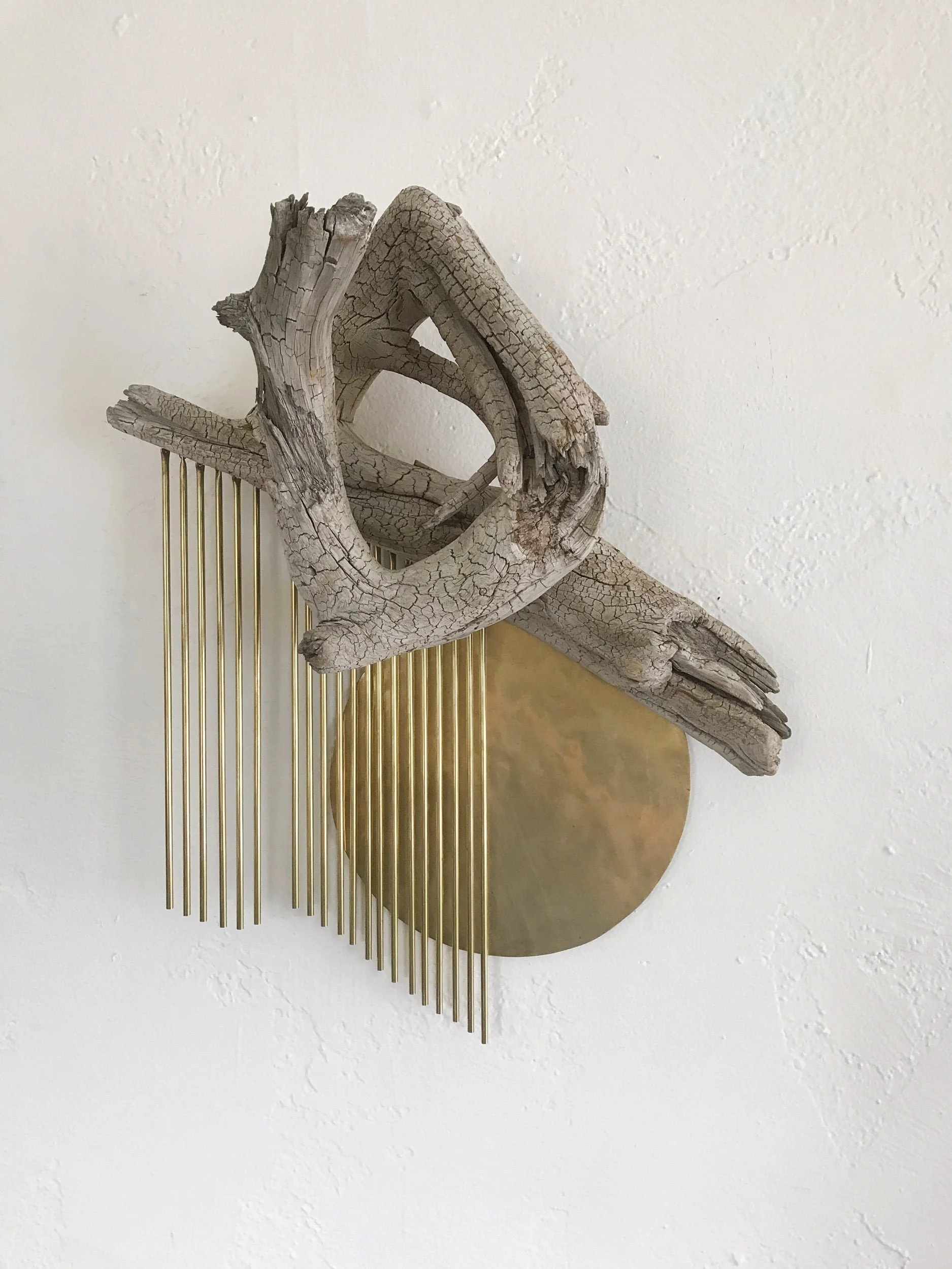 Decorative wall art made of a weathered driftwood piece, a brass sheet circle, and brass metal rods hanging vertically.