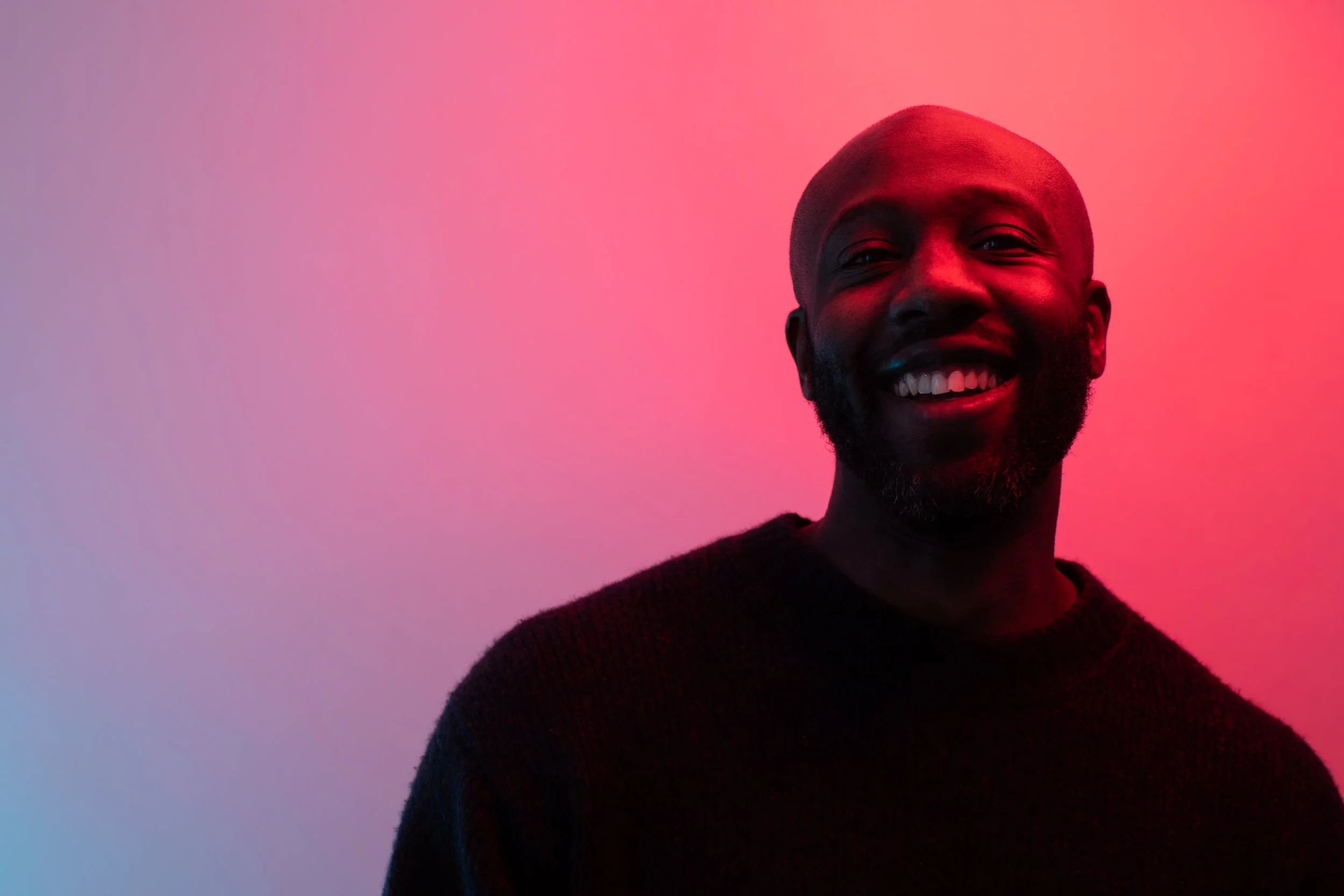 A smiling man with a beard and shaved head, wearing a dark sweater, against a colorful background with pink and blue gradient lighting.