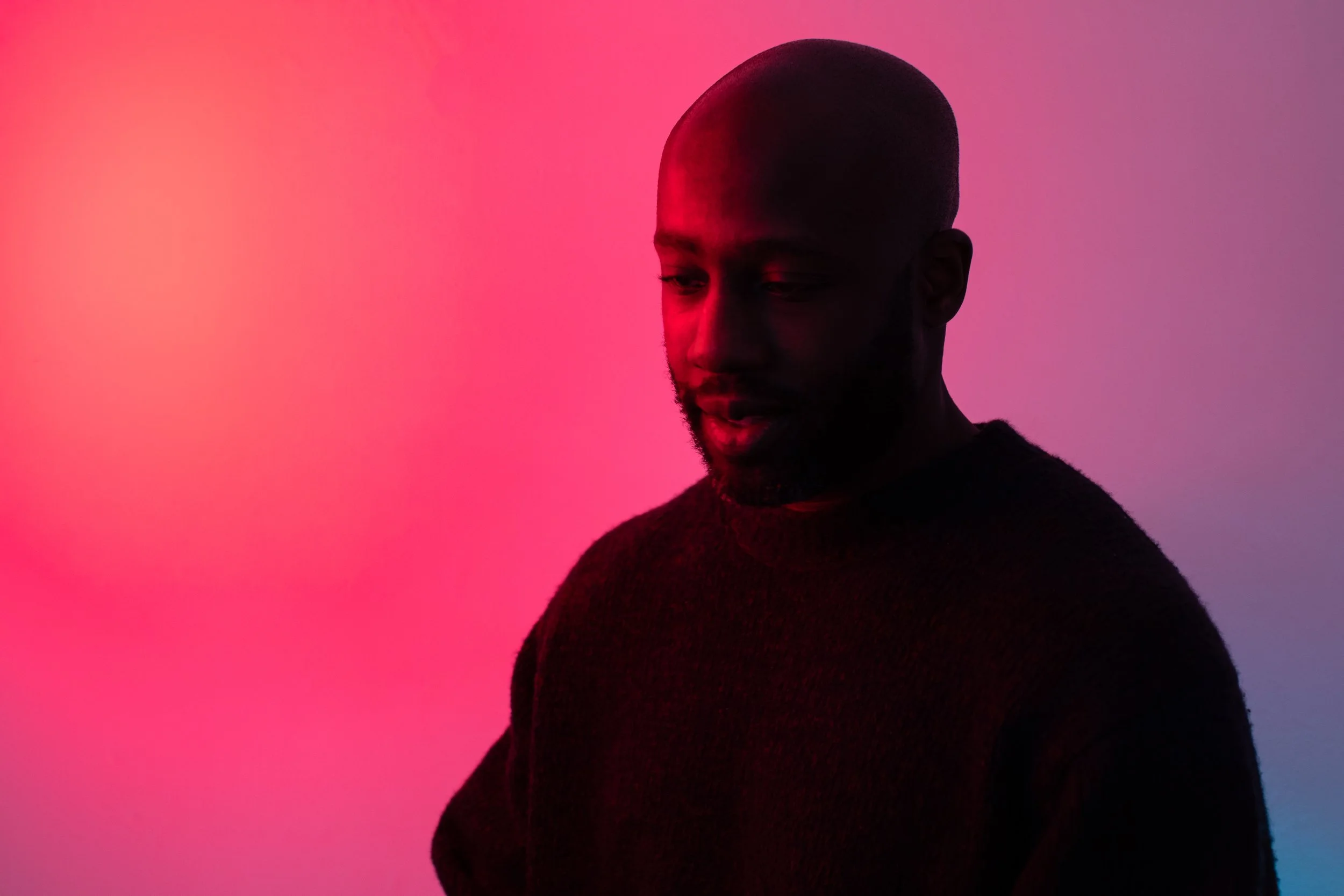 A man with a beard looking down, illuminated by pink and purple lighting, wearing a dark sweater against a gradient pink and purple background.