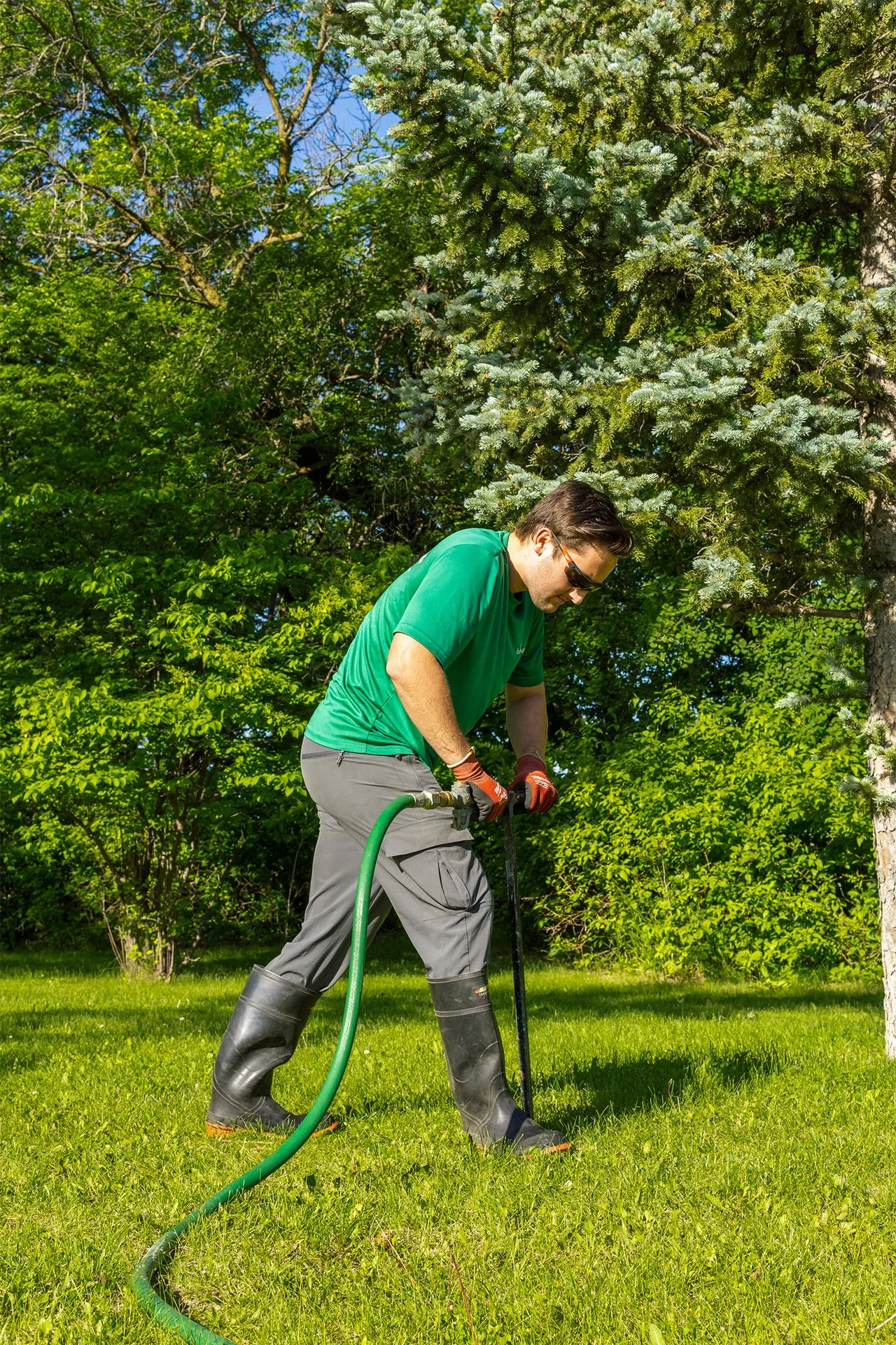 A man in a green shirt and gray pants stands outdoors surrounded by trees and greenery. He is using a tool and wearing gloves and boots, with a green hose nearby.
