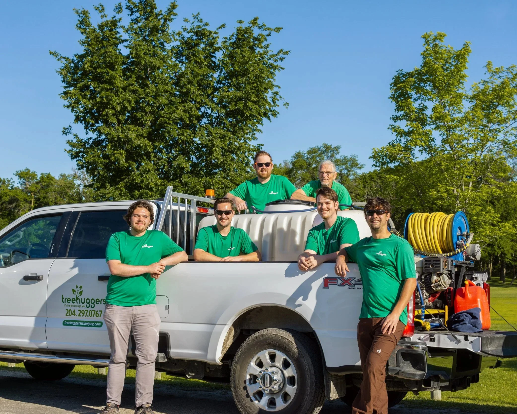 A group of six men in green shirts standing in front of a white truck with equipment mounted on it, including hoses, tools, and a ladder, outdoors under a blue sky with trees.