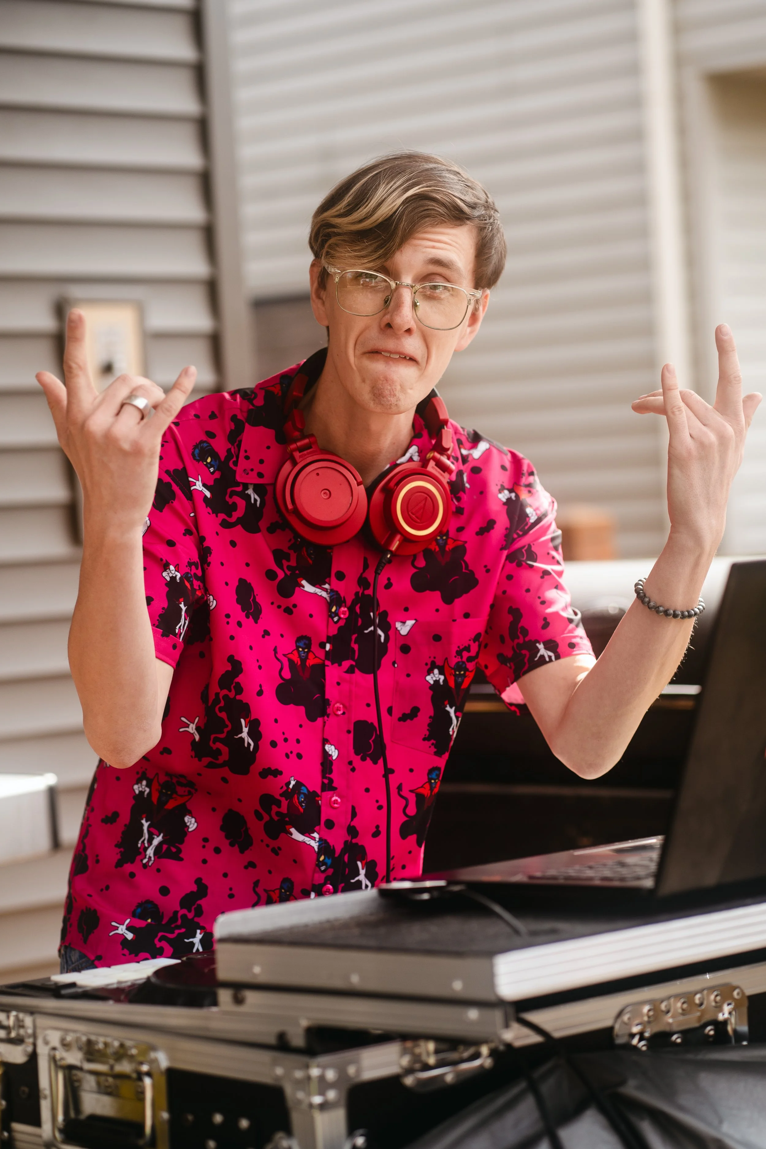 A young man with glasses and worn hair, making a face and gesturing with both hands, wearing a pink and black patterned shirt with large red headphones around his neck, standing behind a DJ turntable setup.