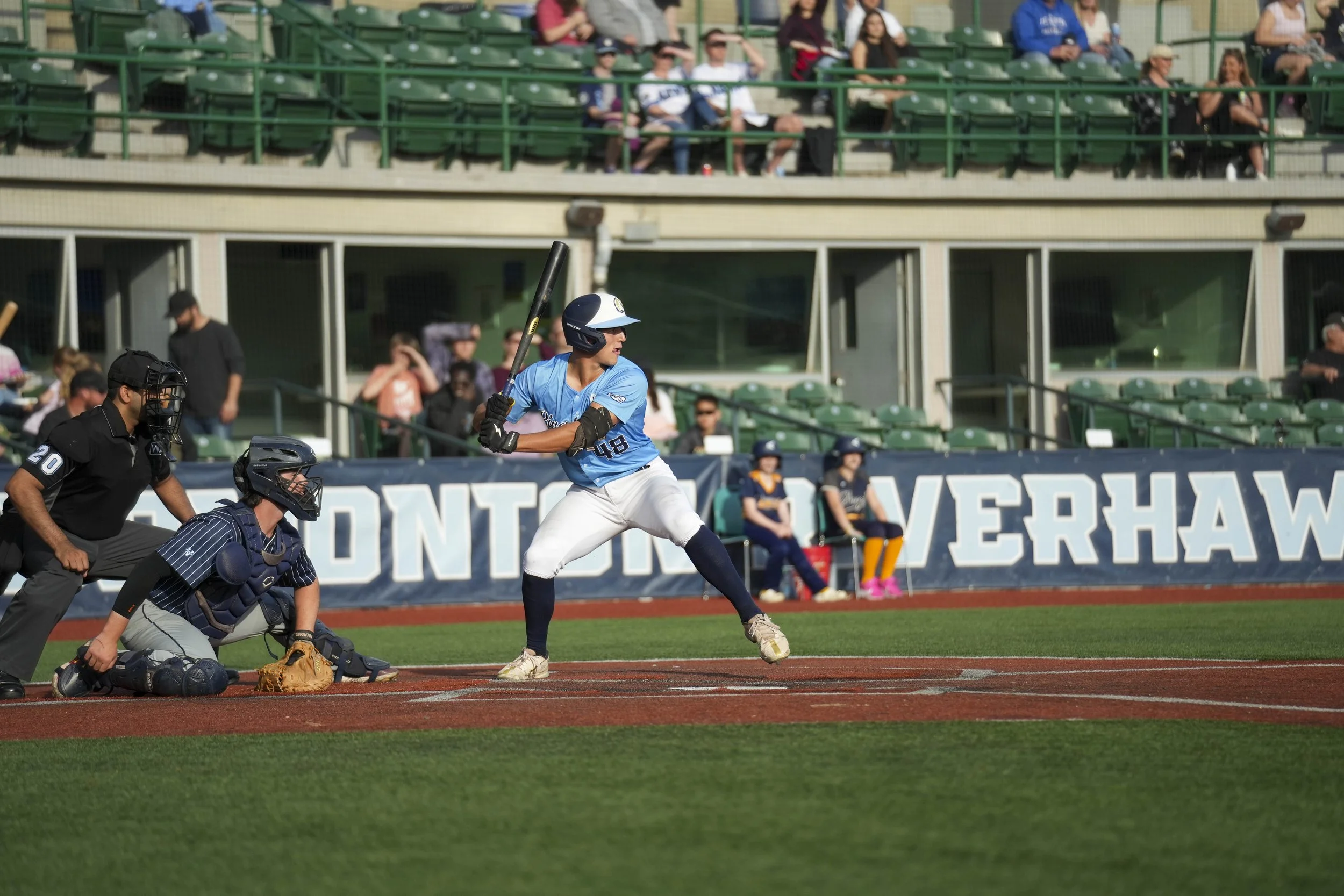 A baseball player in a blue uniform ready to bat at home plate during a game, with an umpire and catcher behind him, and spectators in the stands in the background.