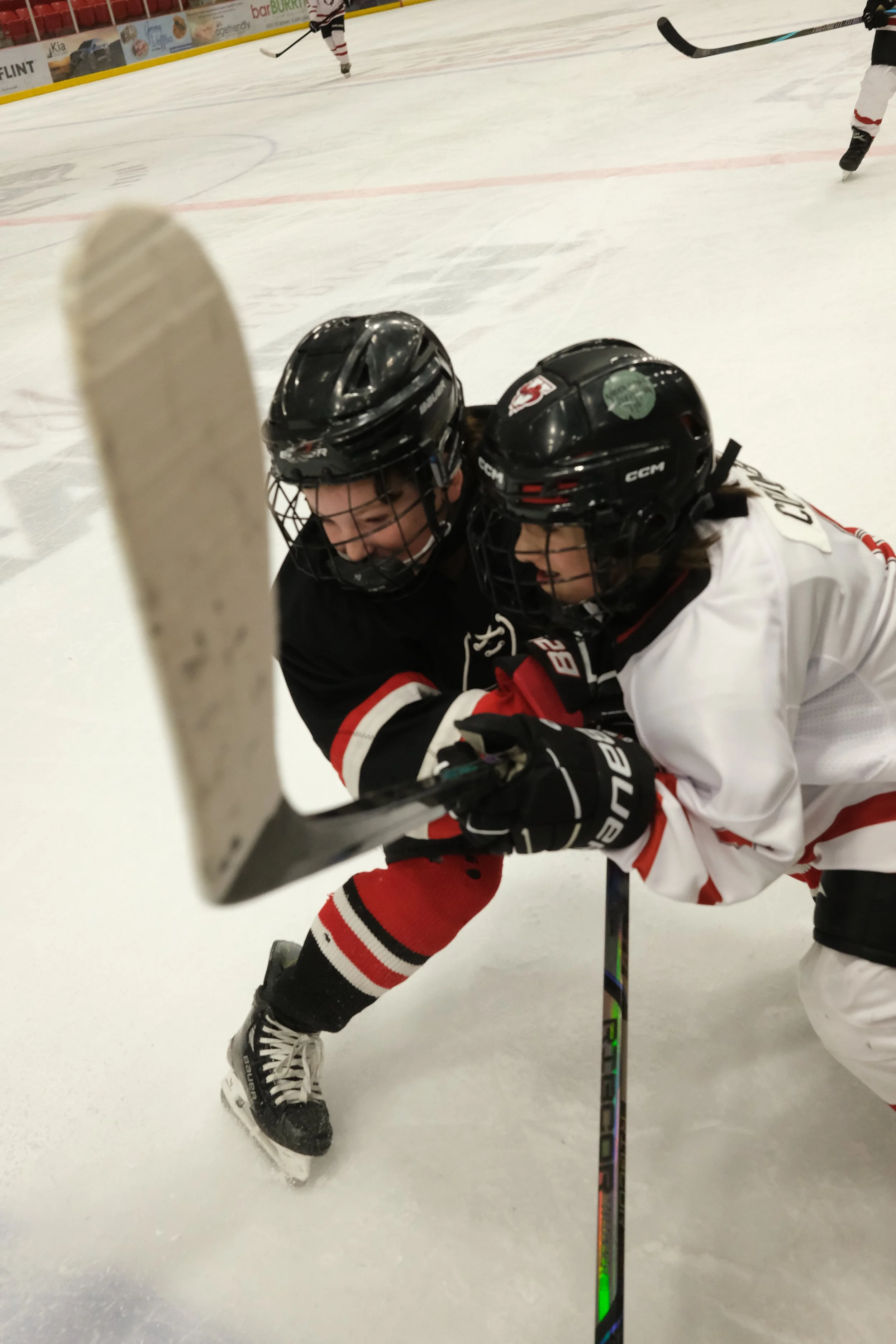 Two ice hockey players, wearing helmets and jerseys, are engaged in a game on the ice rink. One player is lying on the ice while the other appears to be assisting or checking. Hockey sticks and skates are visible, and other players are in the backgro