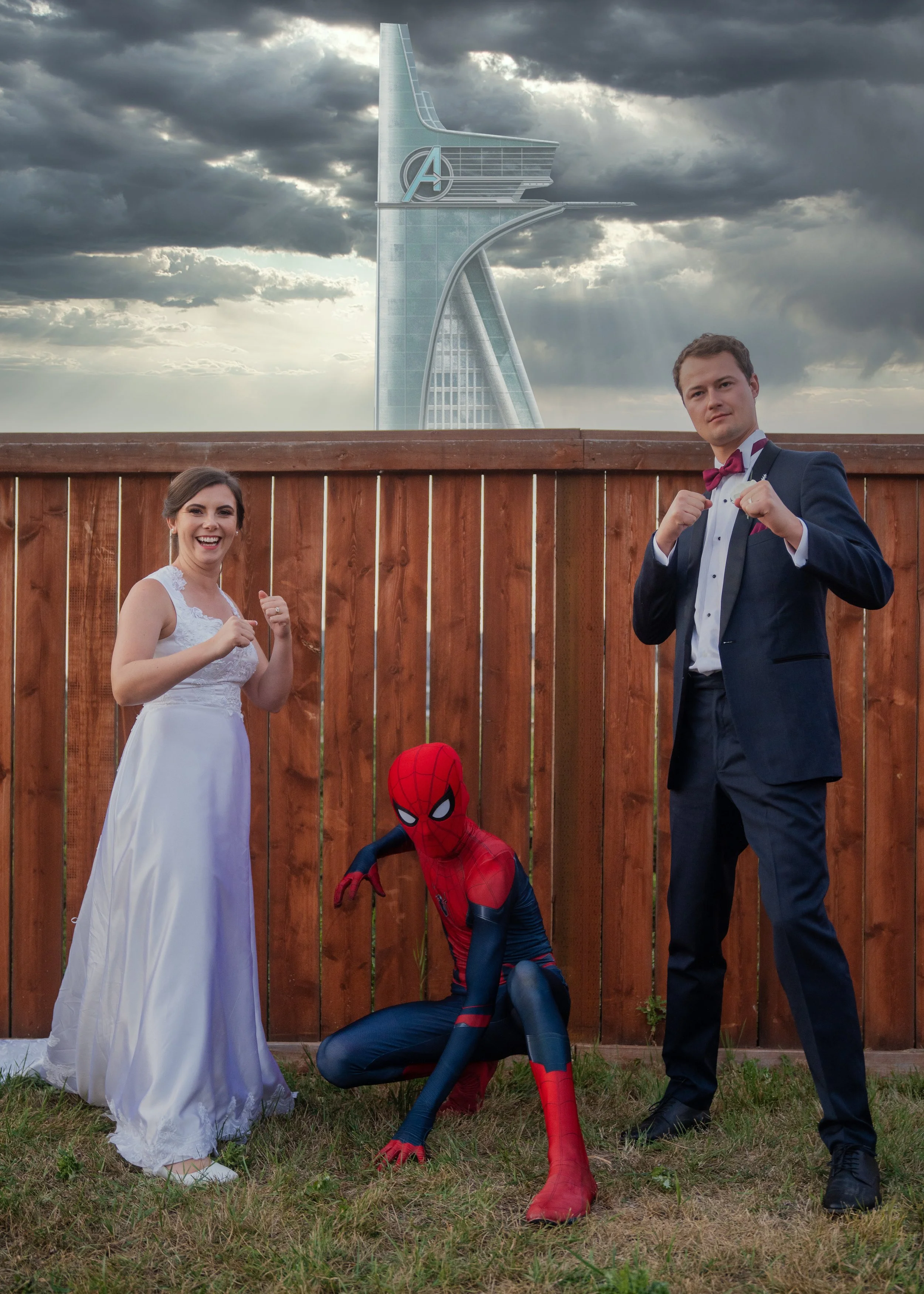 Four people dressed as superheroes, including a woman in a wedding dress, a man in a suit with a bow tie, and a person in a Spider-Man costume, posing in front of a wooden fence and a skyscraper with the Avengers' logo under dark cloudy skies.