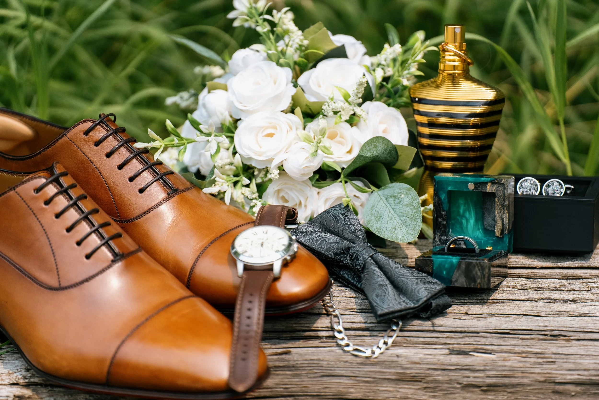 Brown leather dress shoes, a wristwatch, a chain necklace, a floral boutonniere, a perfume bottle with black and gold stripes, cufflinks, and a box with rings are arranged on a rustic wooden surface.