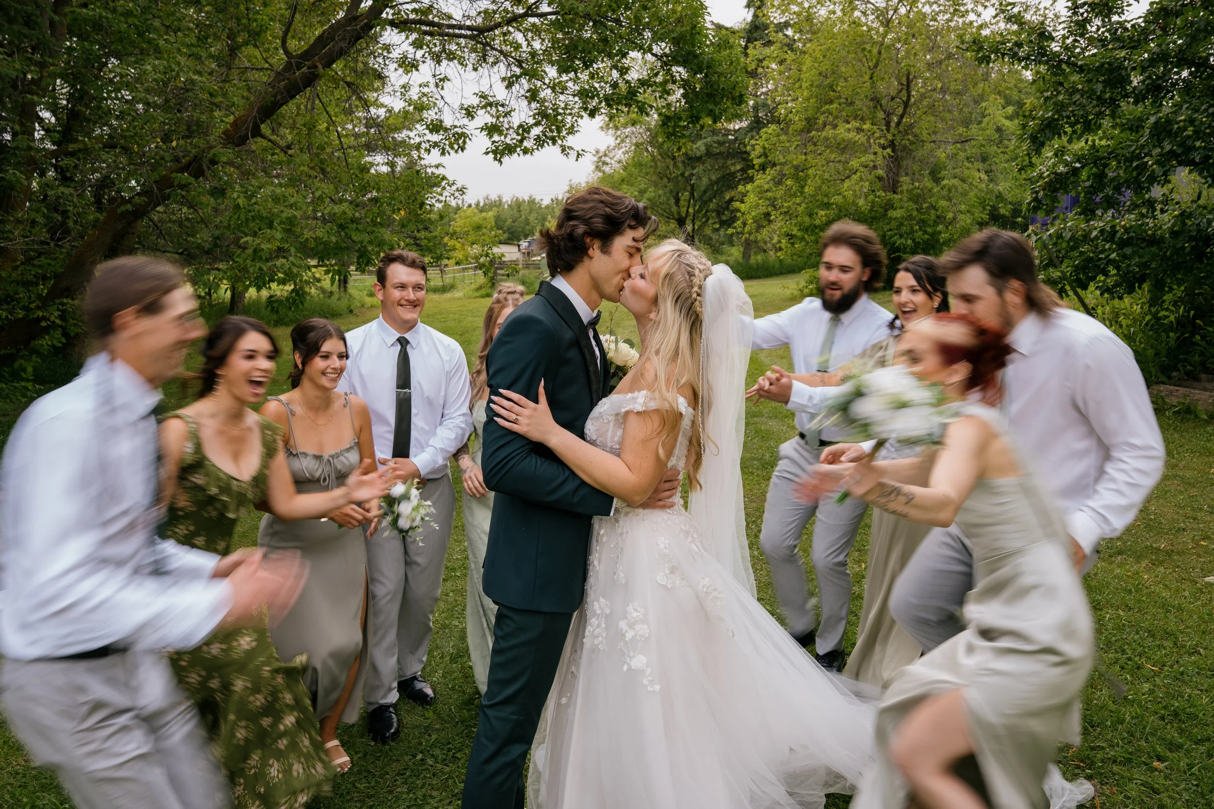 A newlywed couple, a man in a dark suit and a woman in a white wedding dress, kissing in the center of a group of friends celebrating outdoors.