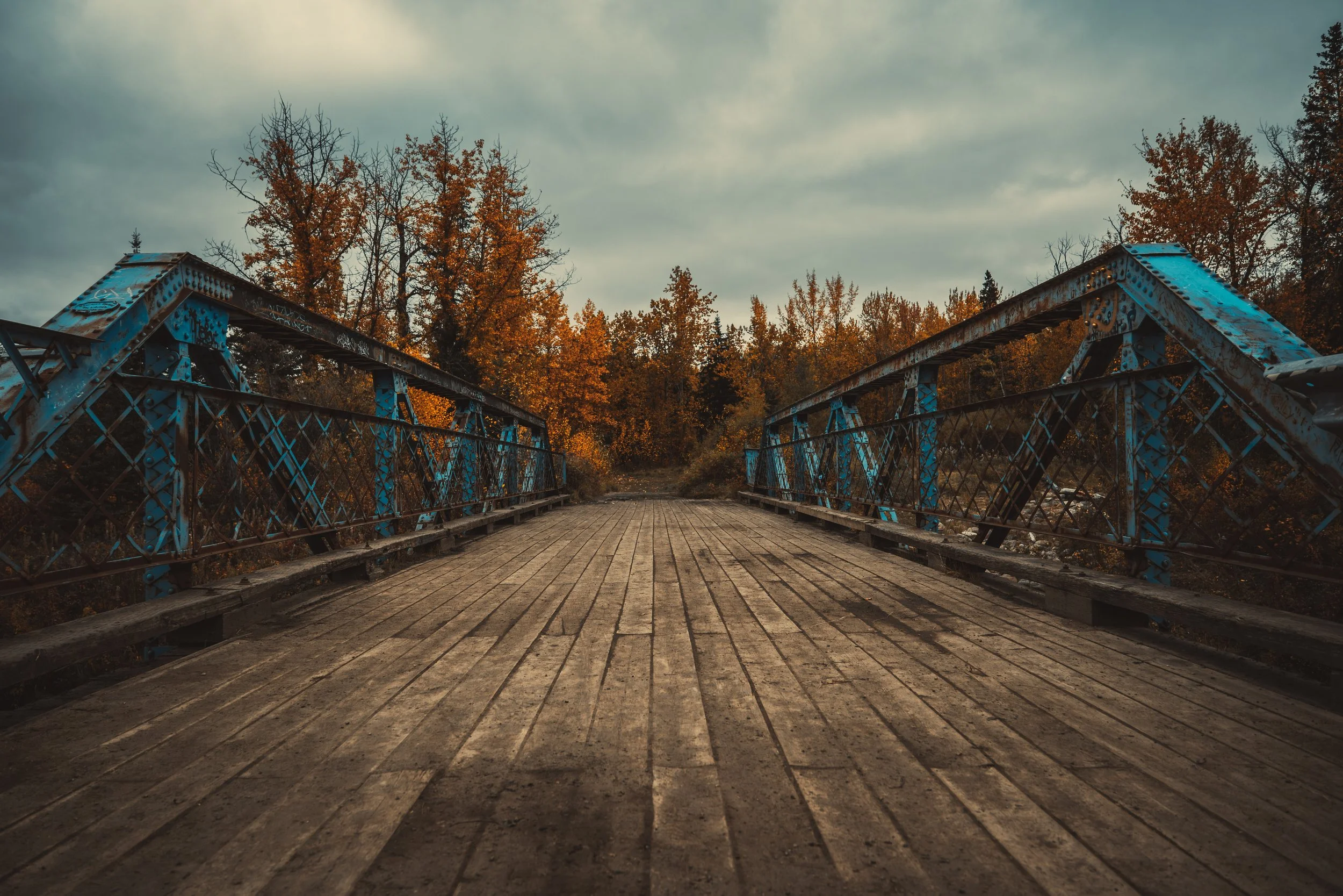 An old blue metal bridge with wooden planks on the walkway, surrounded by trees with autumn foliage and a cloudy sky overhead.