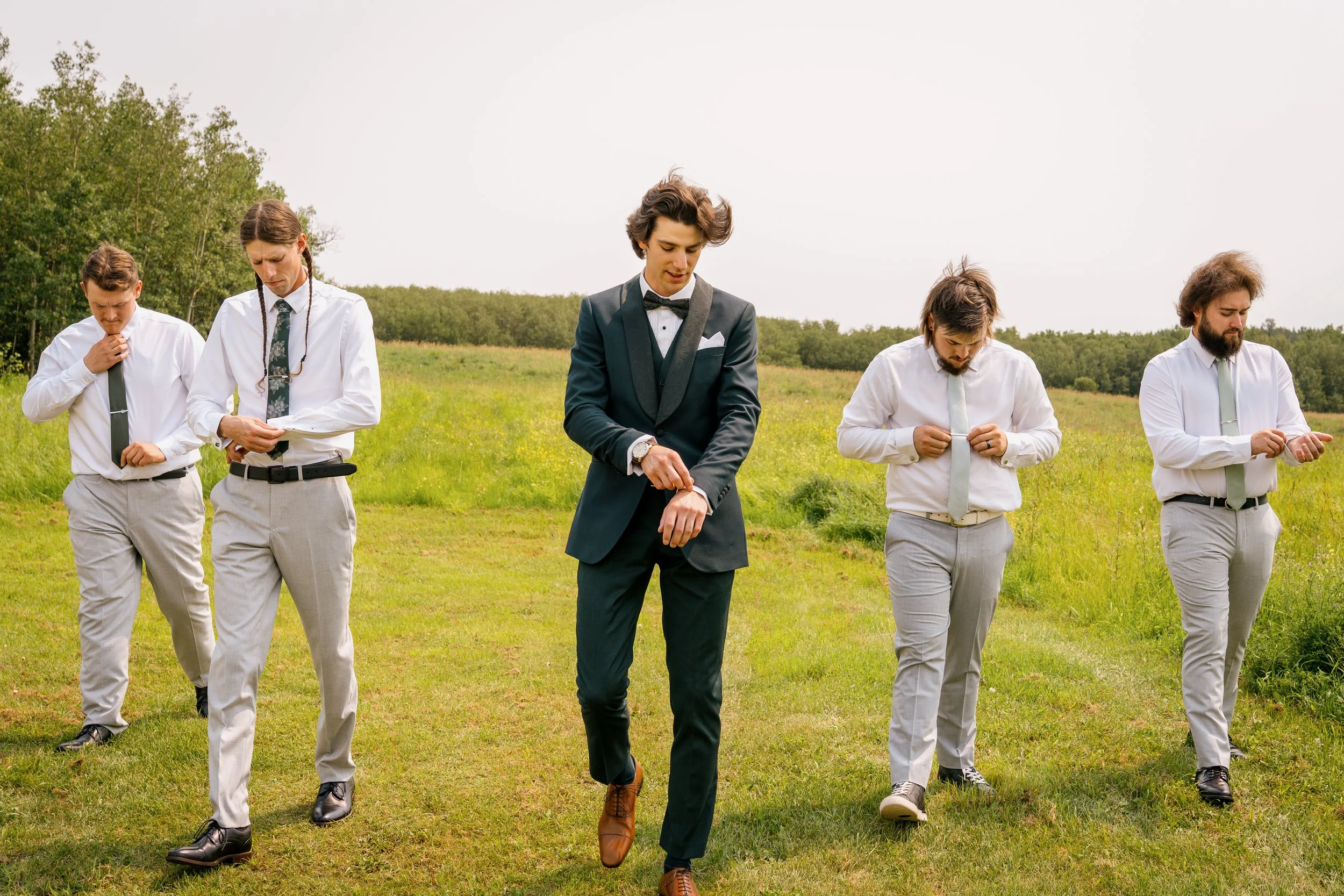 Five men walking in a grassy field, five are dressed in white shirts and ties, one is in a dark suit and bow tie, all looking down as they walk.