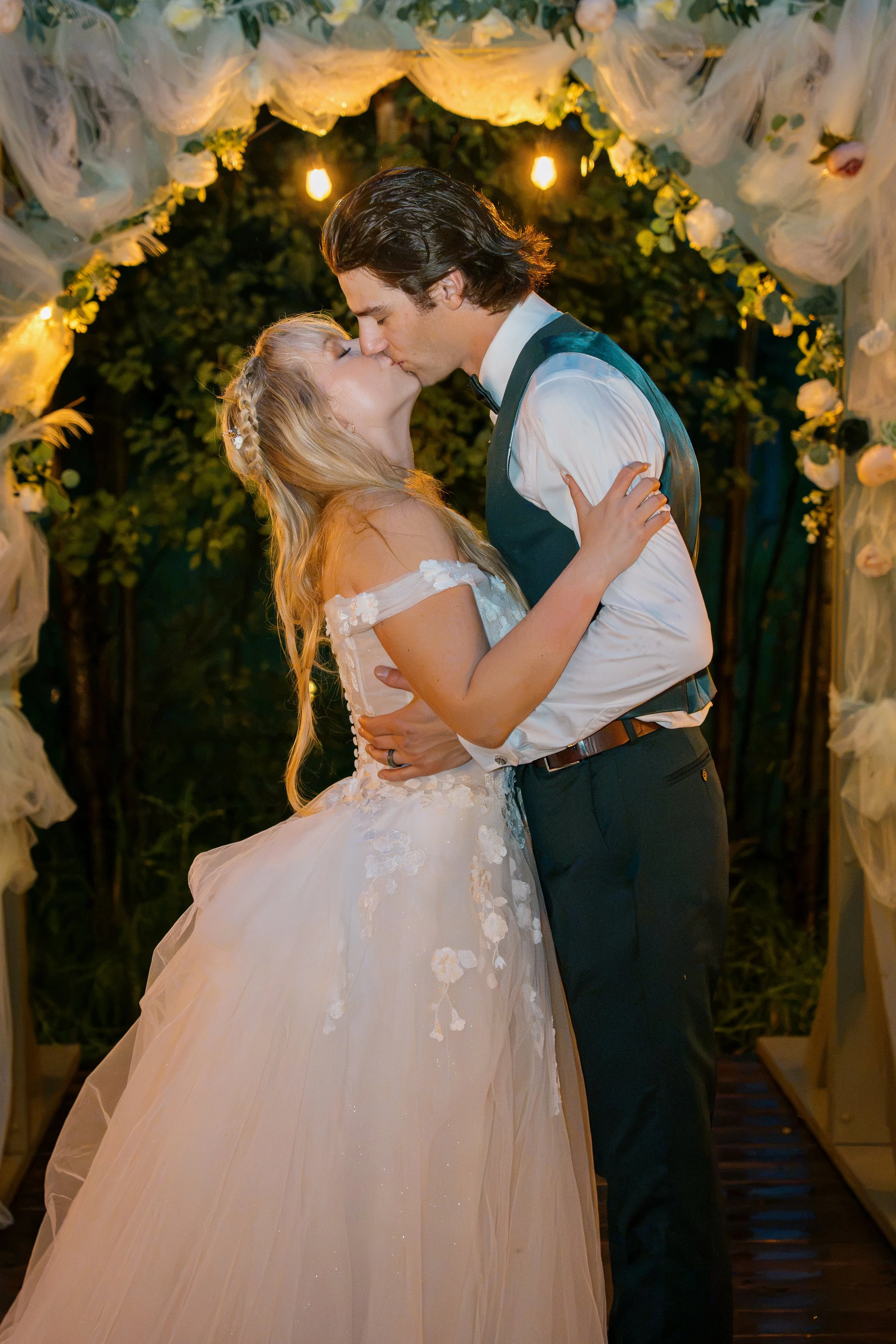 A bride and groom share a kiss under a floral and light decorated arch at their wedding reception.