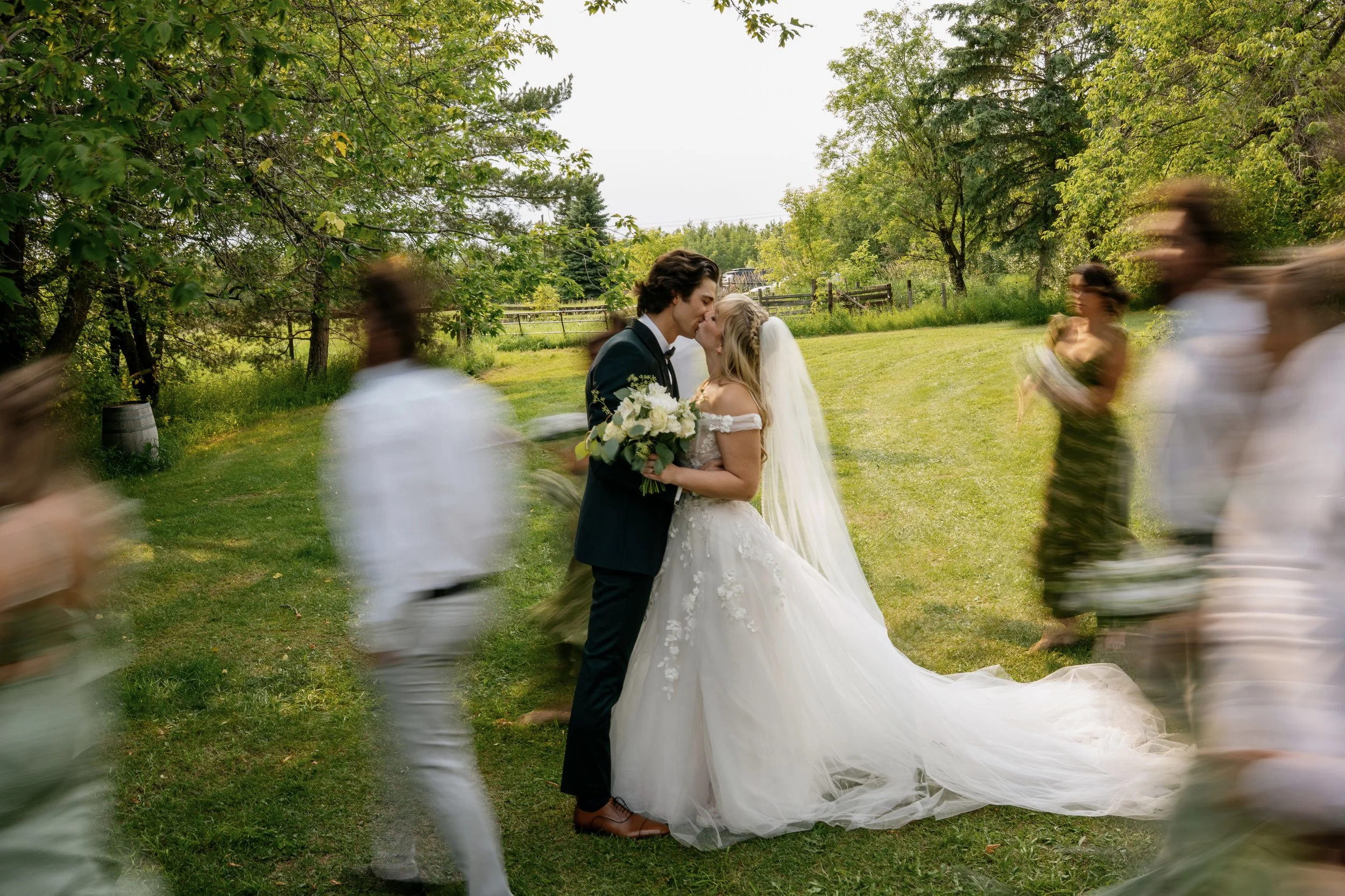 A newlywed couple shares a kiss during their outdoor wedding ceremony with a green, wooded background. Guests are walking or dancing around them, appearing blurred.
