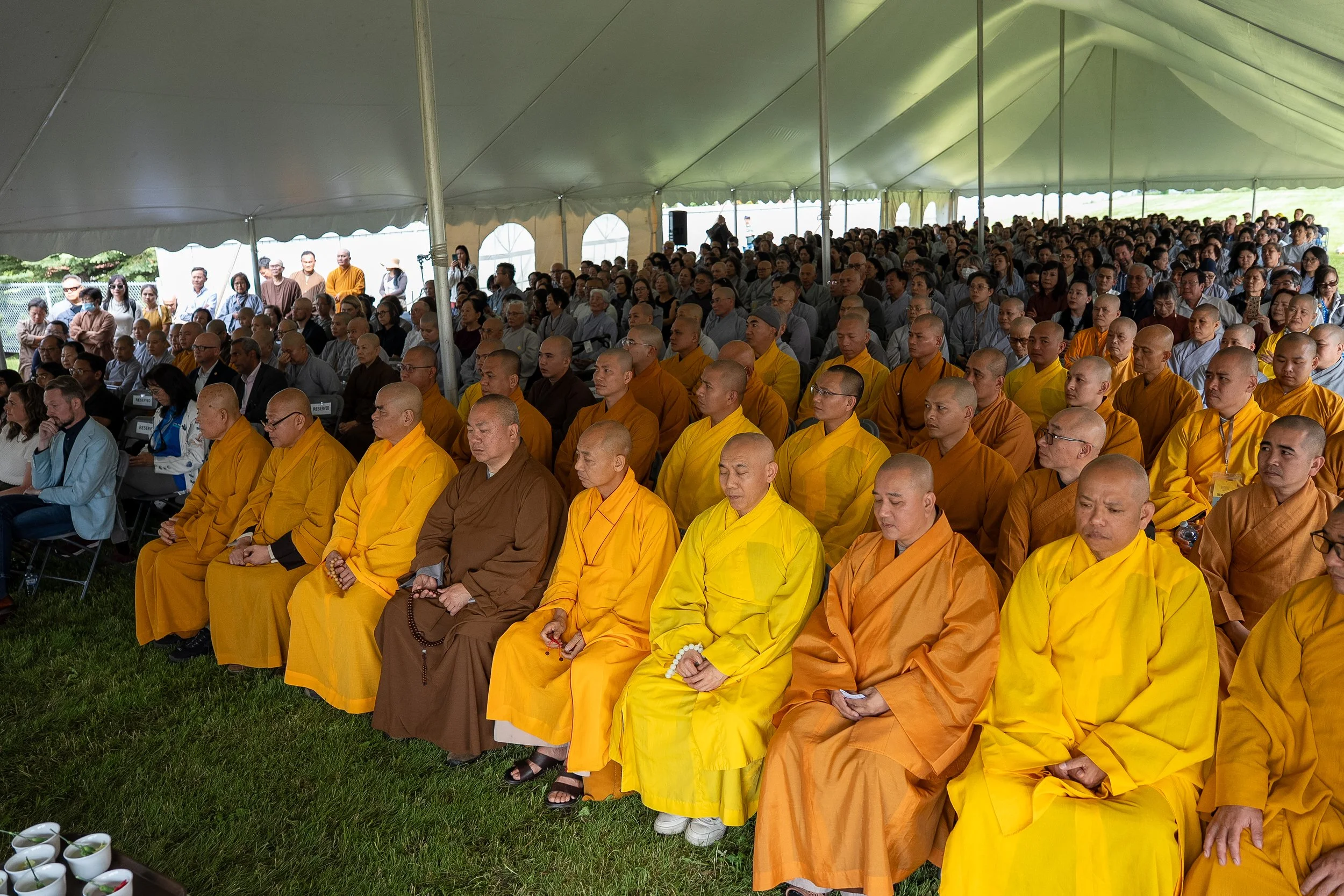 A large gathering of Buddhist monks in yellow robes and other attendees seated under a large tent during an outdoor event.