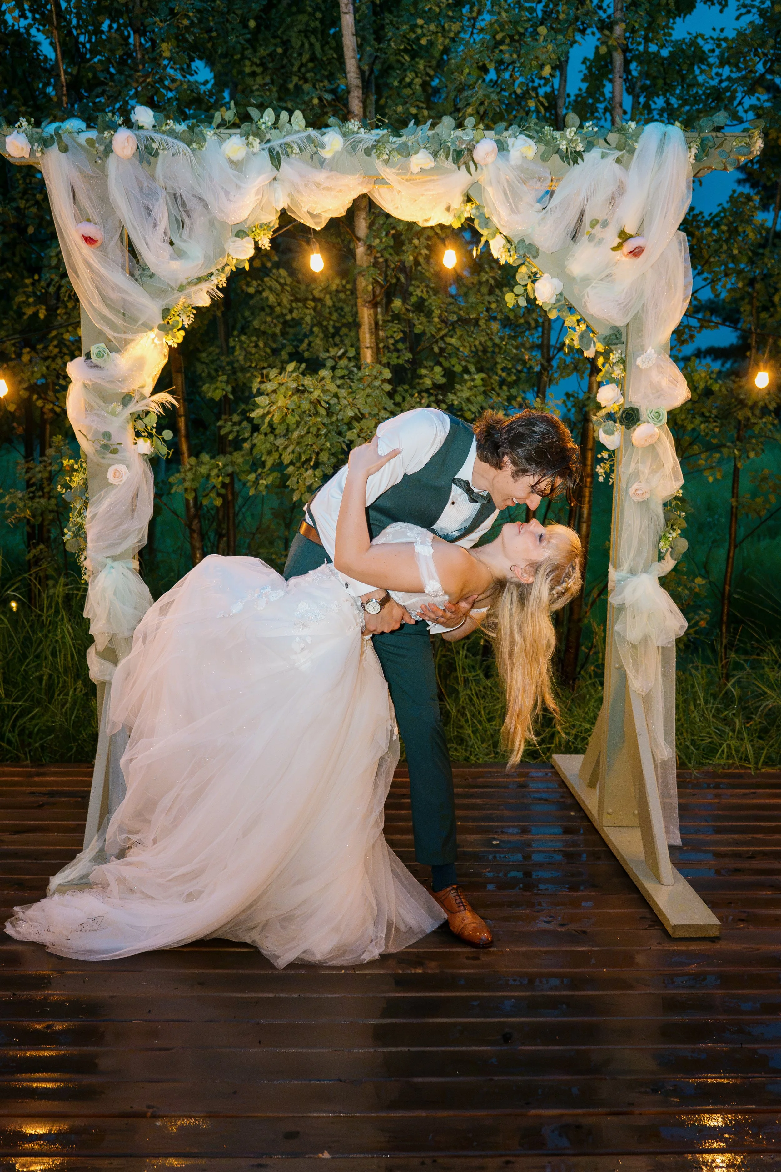 A bride and groom dance under a floral and tulle arch at an outdoor wedding during evening, surrounded by string lights and trees.