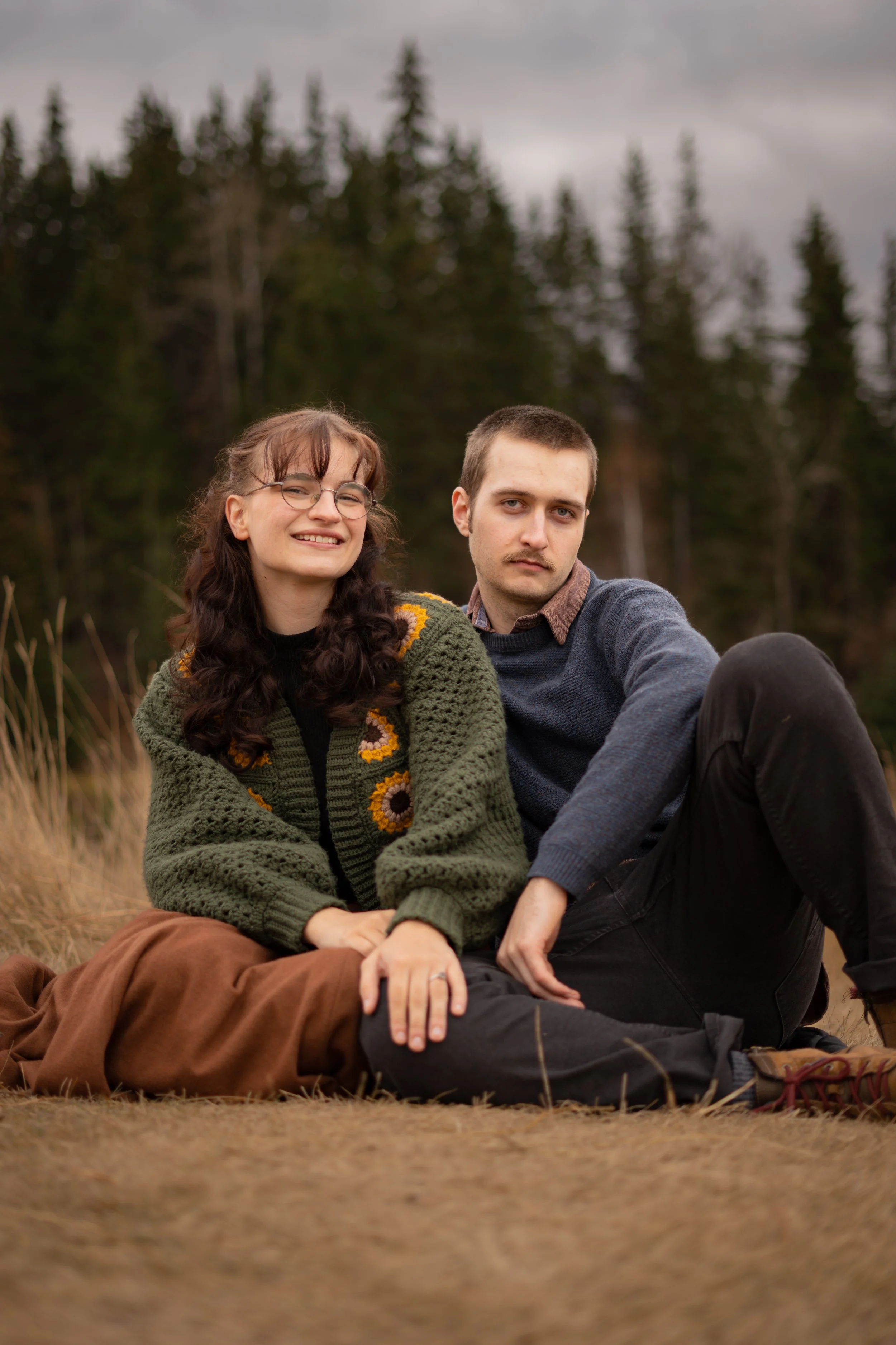 A young woman and a young man sitting outdoors on dry grass with a forest of tall trees in the background. The woman is smiling and wearing glasses, a green knitted sweater, and brown pants. The man has a neutral expression, short hair, a brown shirt