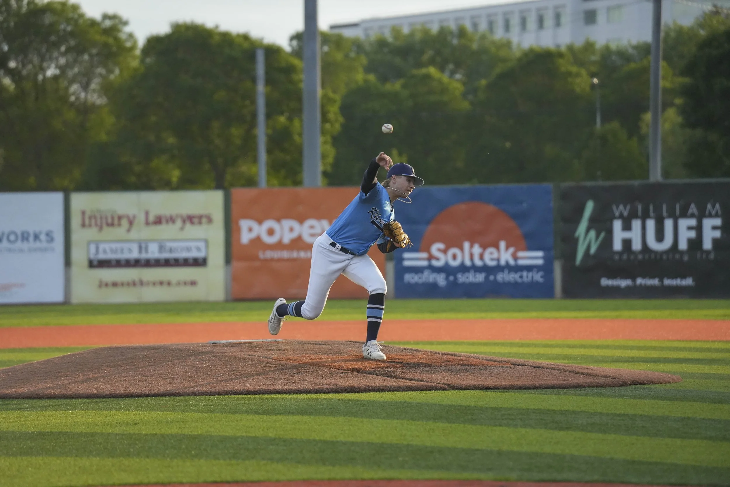 A baseball player in a blue jersey and white pants is pitching the ball on a baseball field during daylight. The player is in a dynamic pose, with one leg raised and arm extended forward. The background features trees and advertisements on a fence.