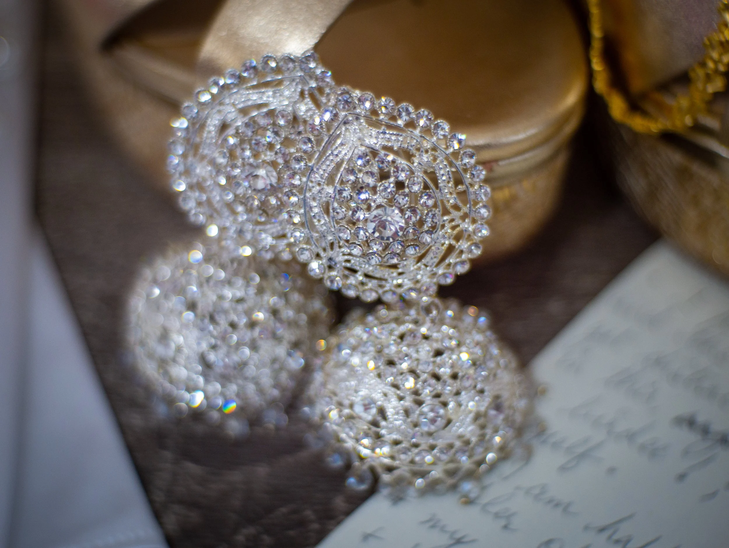 A close-up of a silver jewelry piece with numerous small clear stones, placed on a background that looks like a dark fabric or paper with handwritten text, surrounded by gold-colored objects.