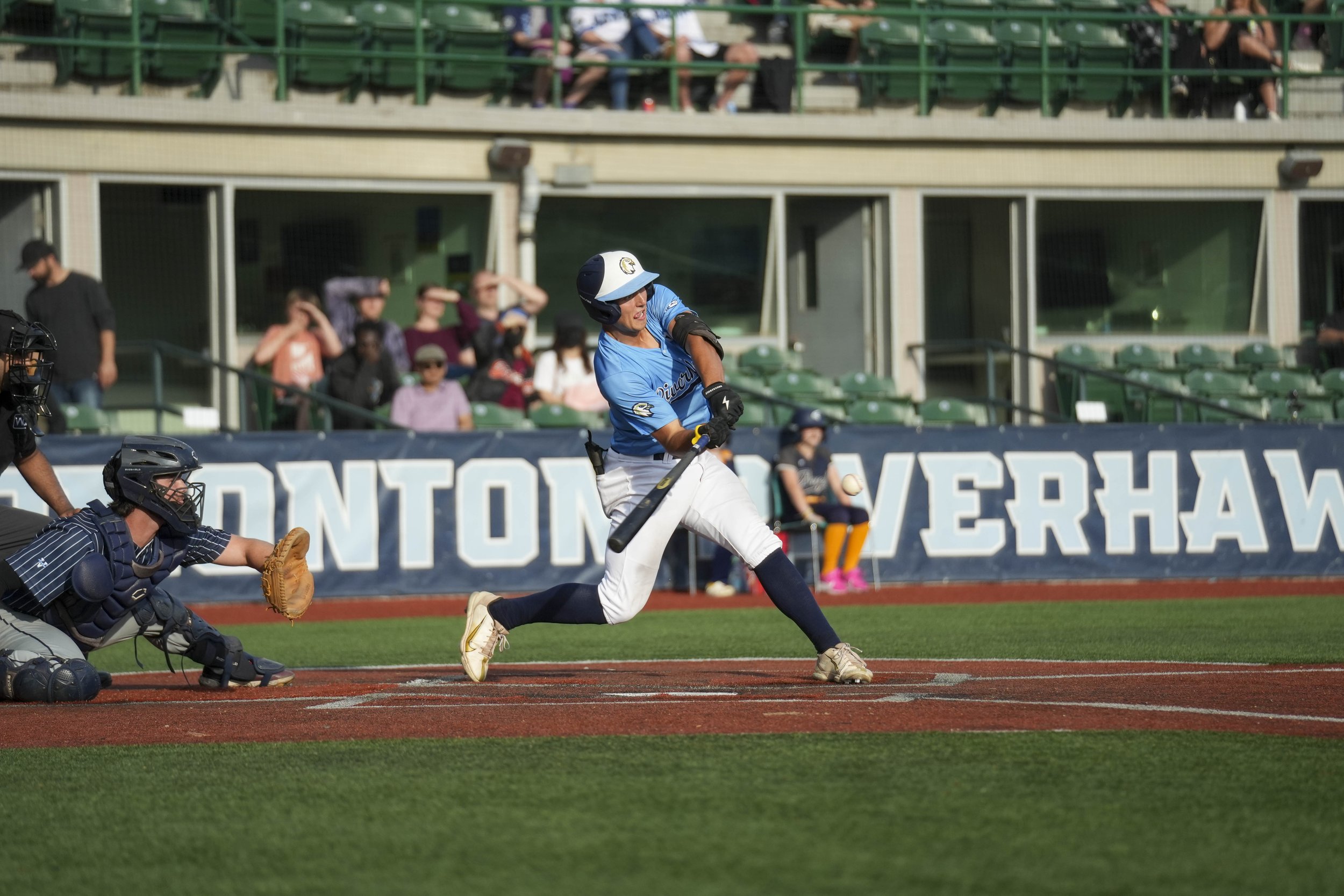 A baseball player in a blue uniform and helmet is batting at a baseball game, with the catcher in black protective gear behind him and spectators in the background.