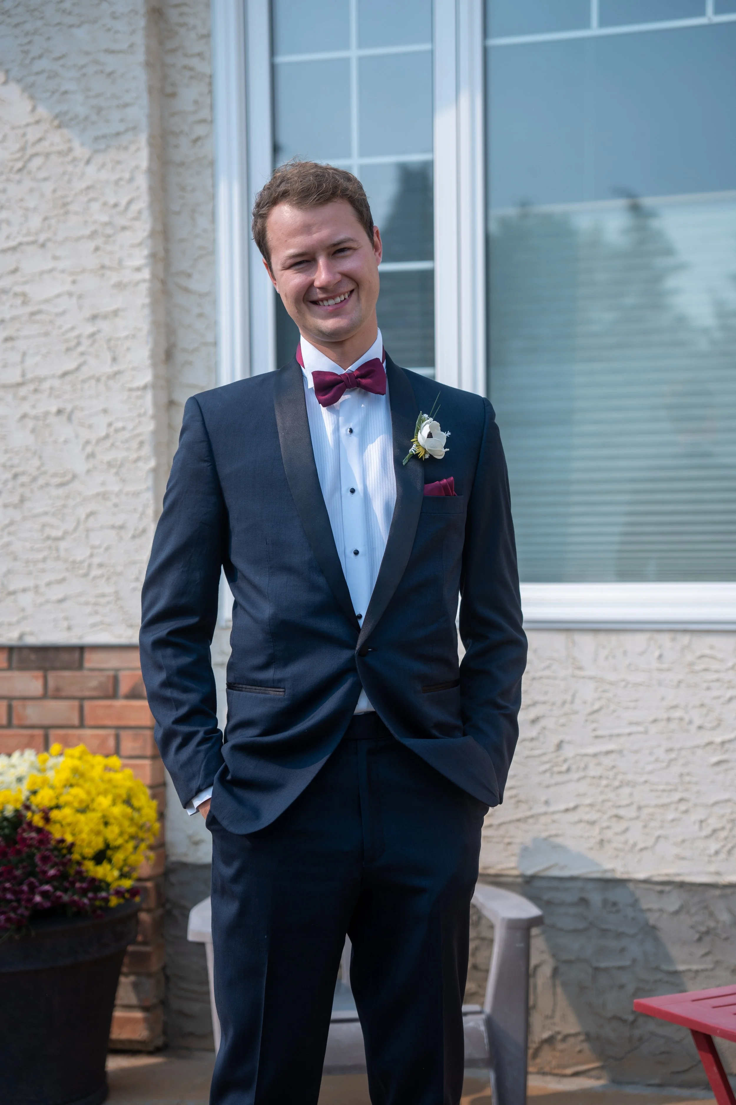 A young man in a formal tuxedo with a burgundy bow tie and boutonniere, smiling outdoors in front of a house with a window and brick exterior, potted flowers, and outdoor chairs.