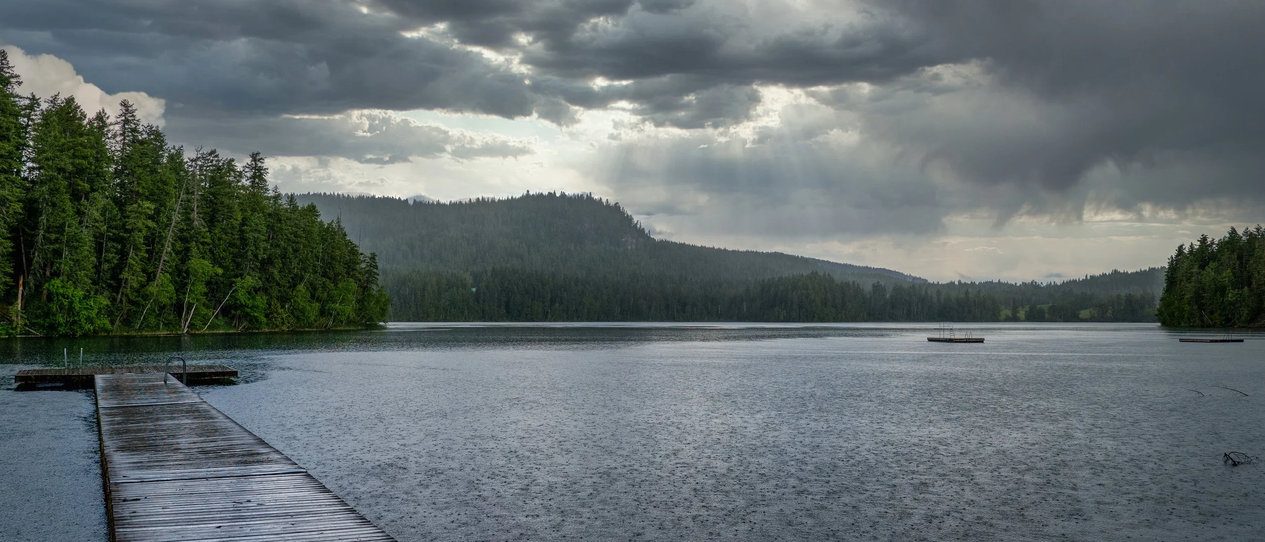 A wooden dock extends into a calm lake surrounded by dense pine trees under a cloudy sky with sunlight breaking through.