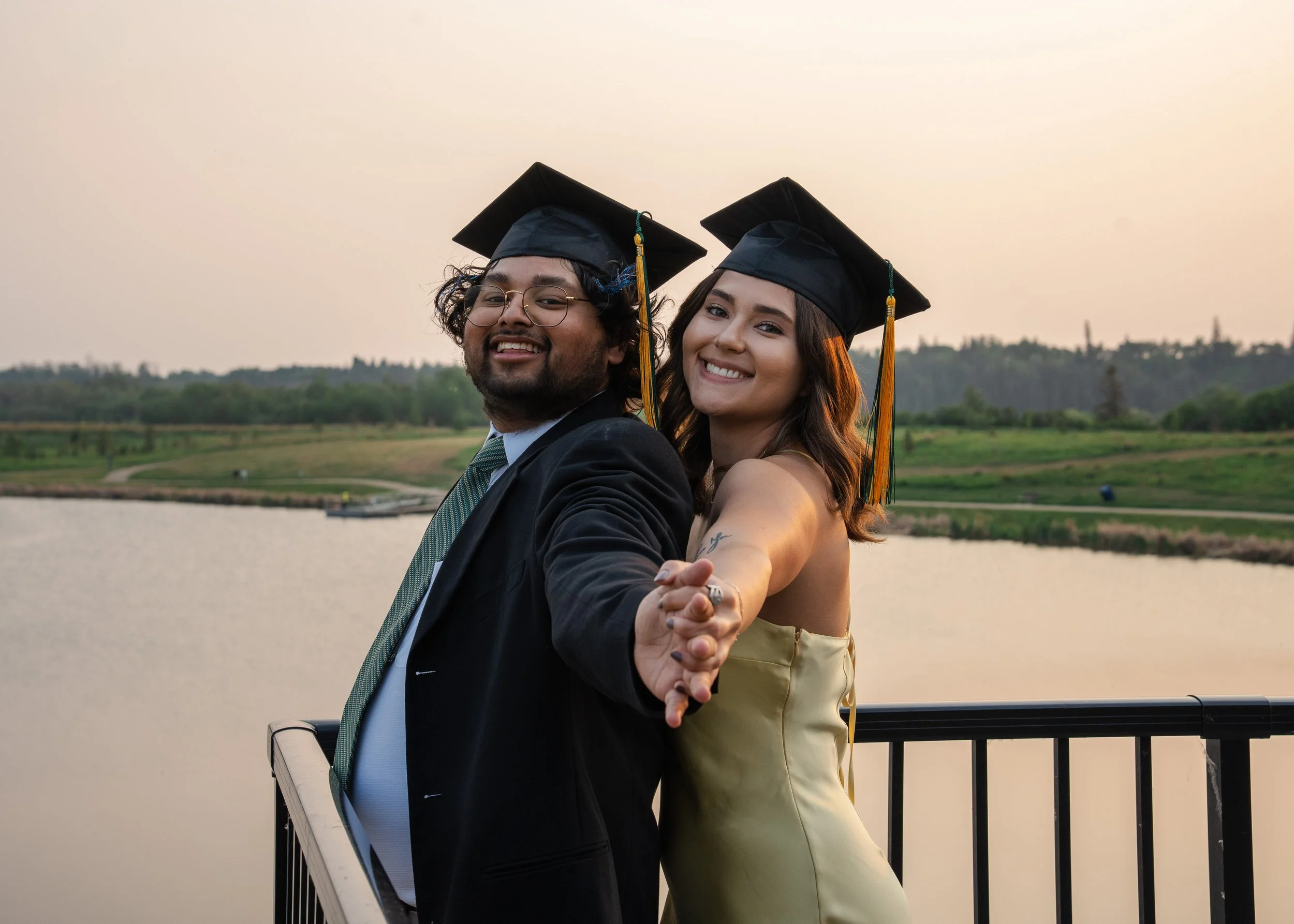Two graduates, a man and a woman, wearing black caps and gowns, are taking a selfie outdoors by a body of water during sunset. They are smiling and appear happy.