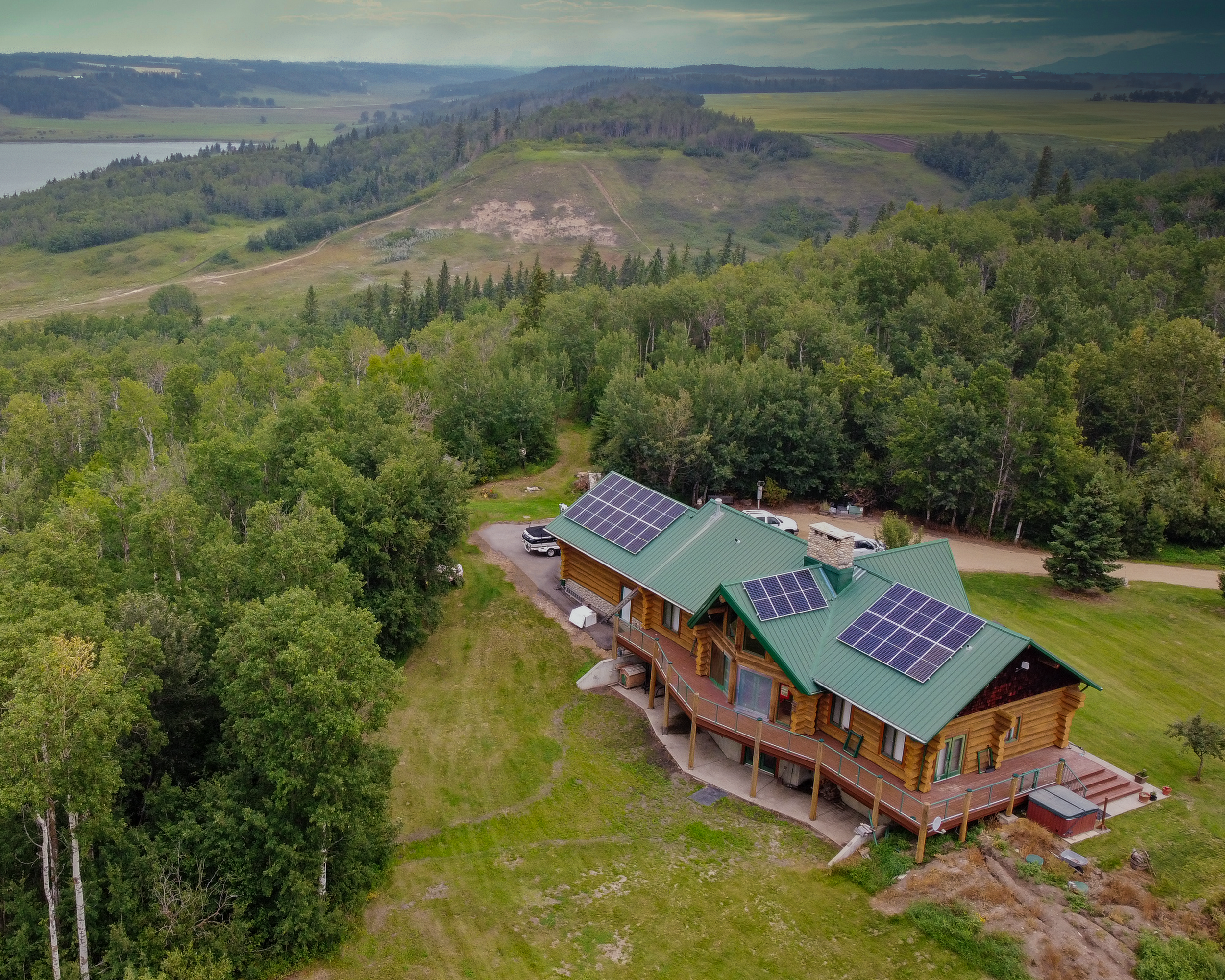Aerial view of a house with solar panels on the roof, surrounded by green trees and forest, with hills and water in the background.
