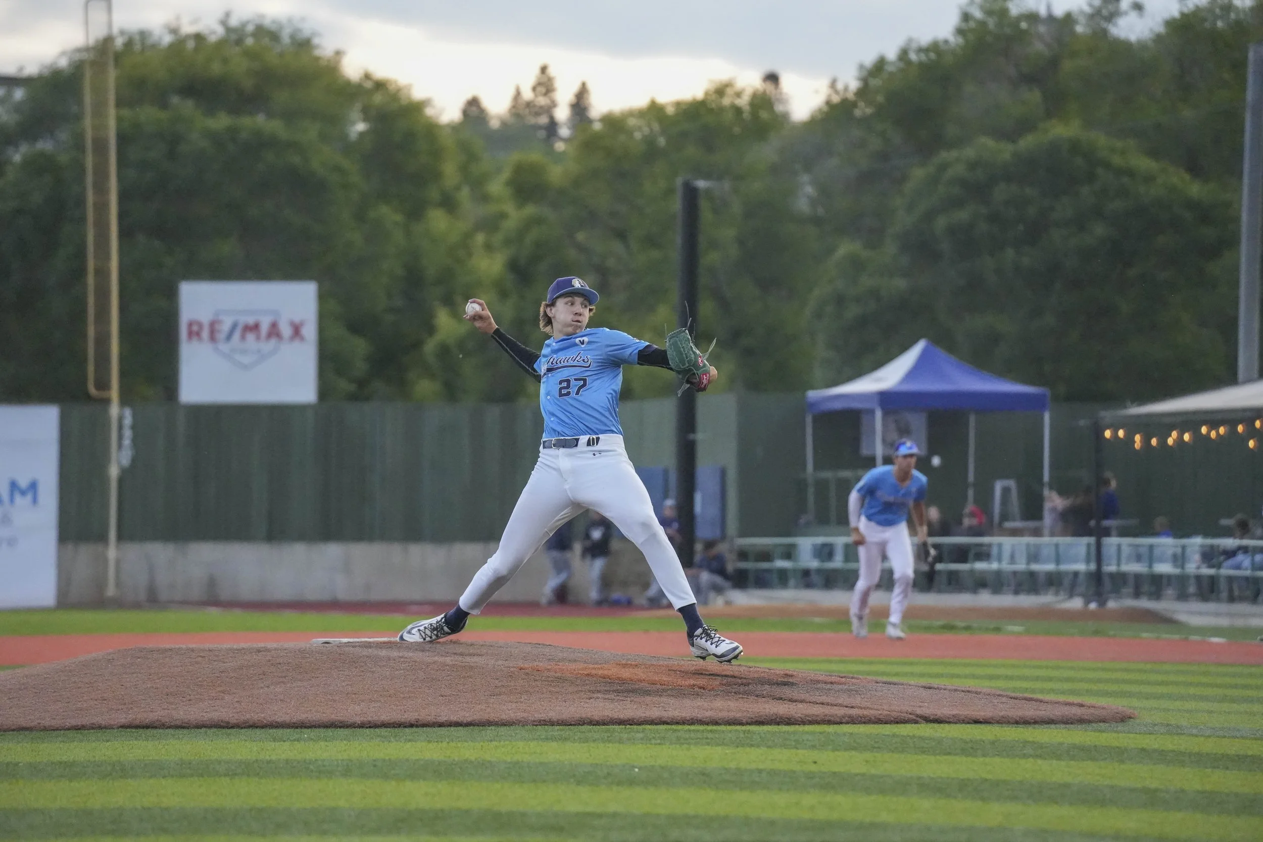 A young male baseball player in a blue jersey and white pants is pitching on the mound during a game, with a green glove on his left hand and a baseball in his right hand, on a baseball field with trees and signs in the background.