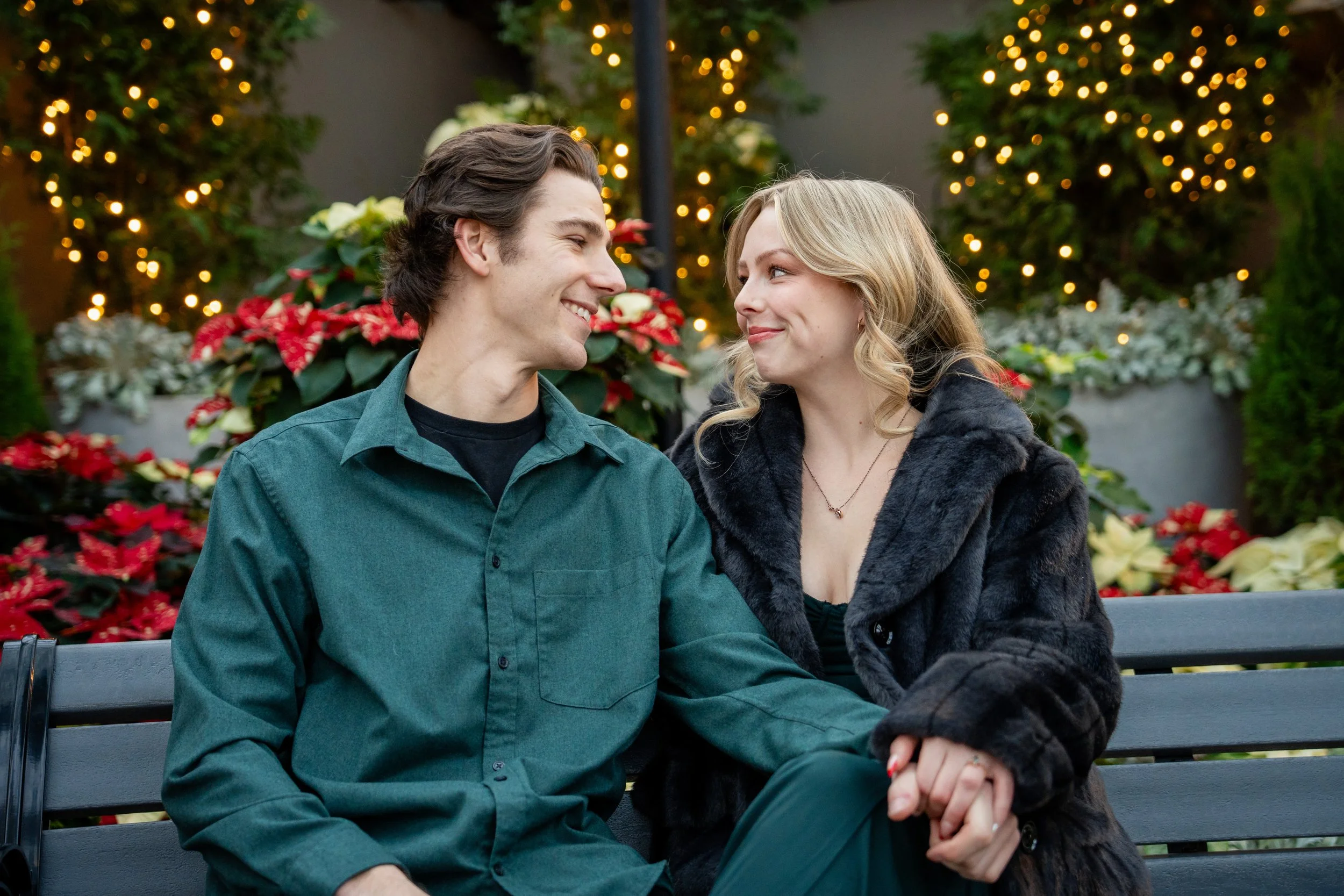 A smiling man and woman sitting closely on a park bench, looking at each other with affection. They are surrounded by festive holiday decorations including poinsettias and blurred string lights in the background.
