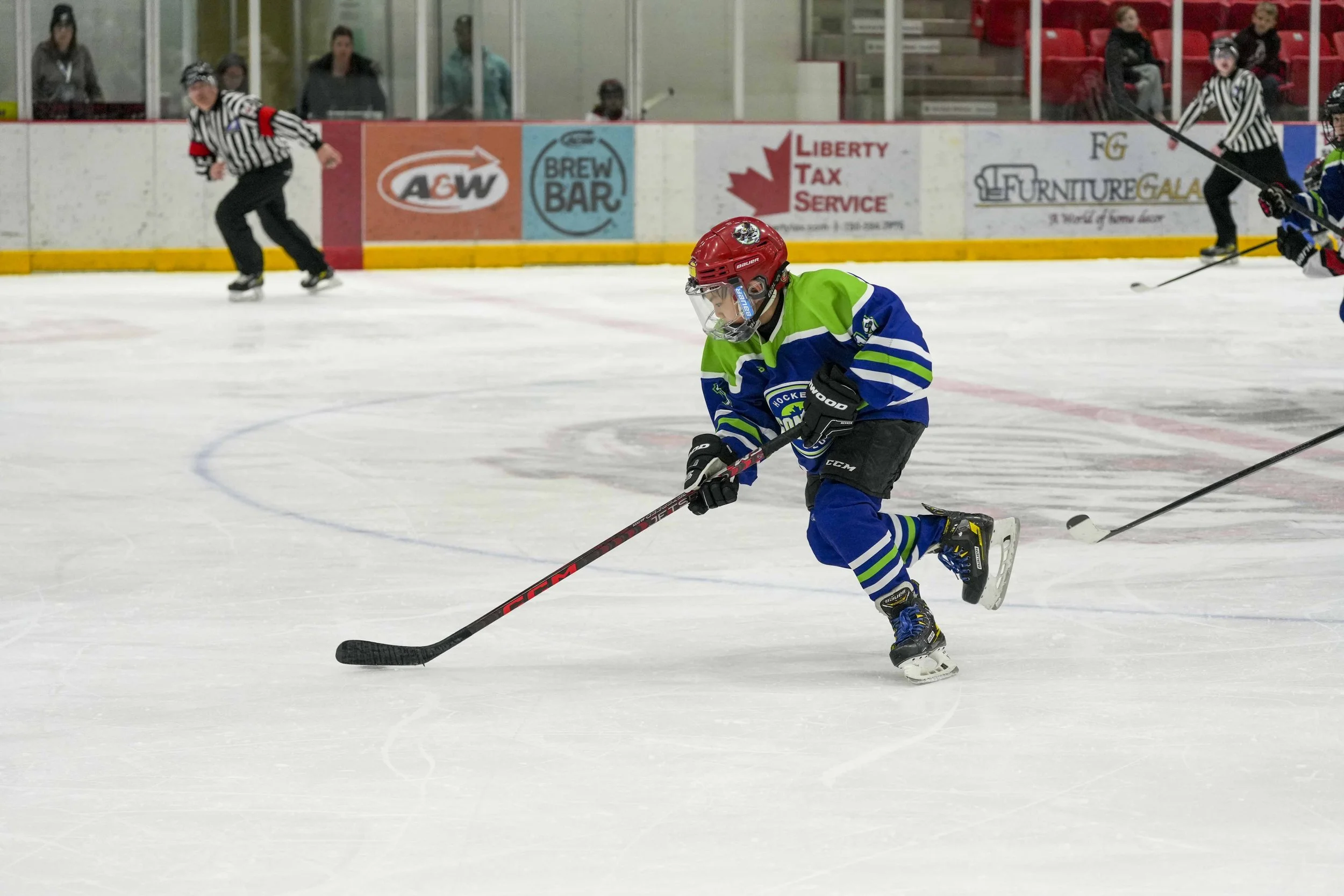 A young hockey player in a green and blue jersey skating on the ice with a hockey stick, wearing a red helmet and black gloves. There are officials and spectators in the background at the ice rink.