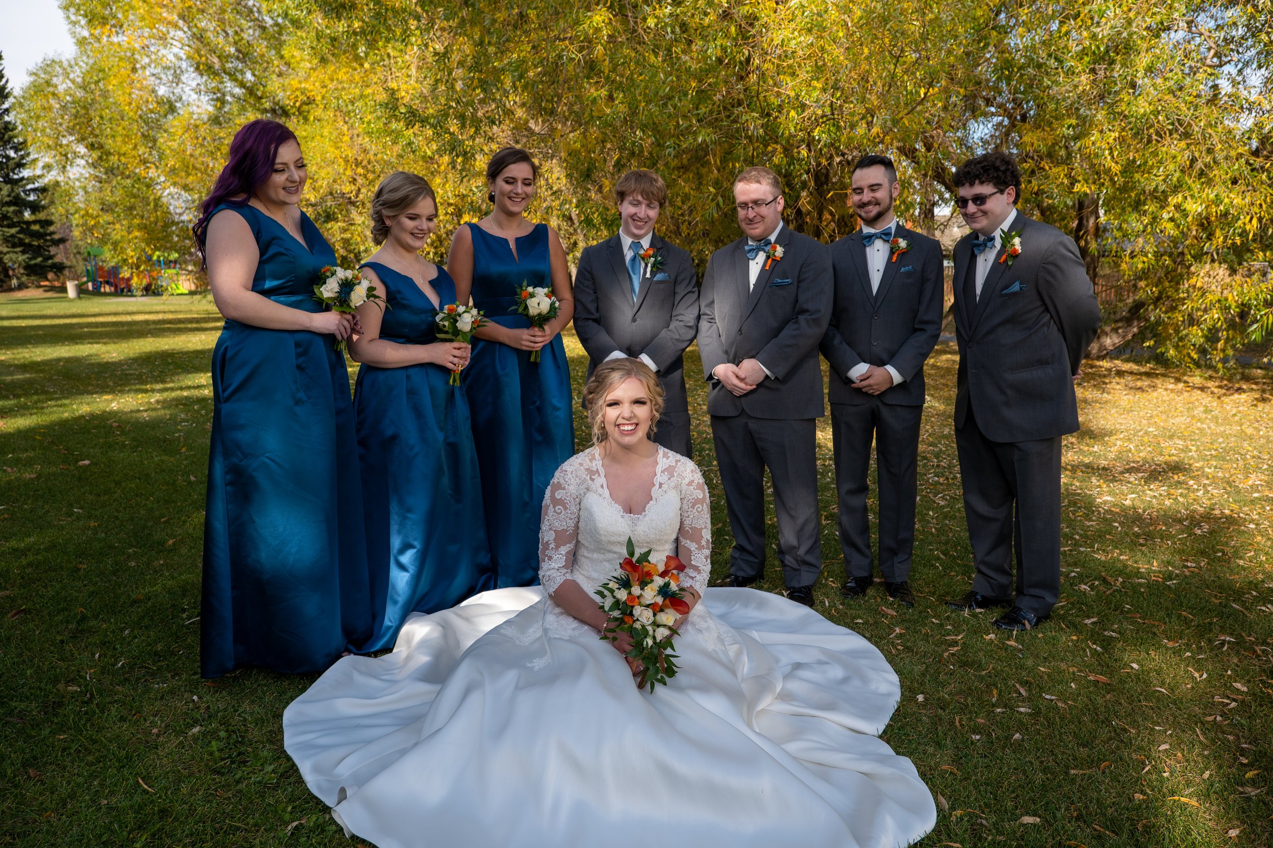 A bride sitting on the ground holding a bouquet, surrounded by eight bridesmaids and groomsmen standing outdoors in a park with autumn foliage.