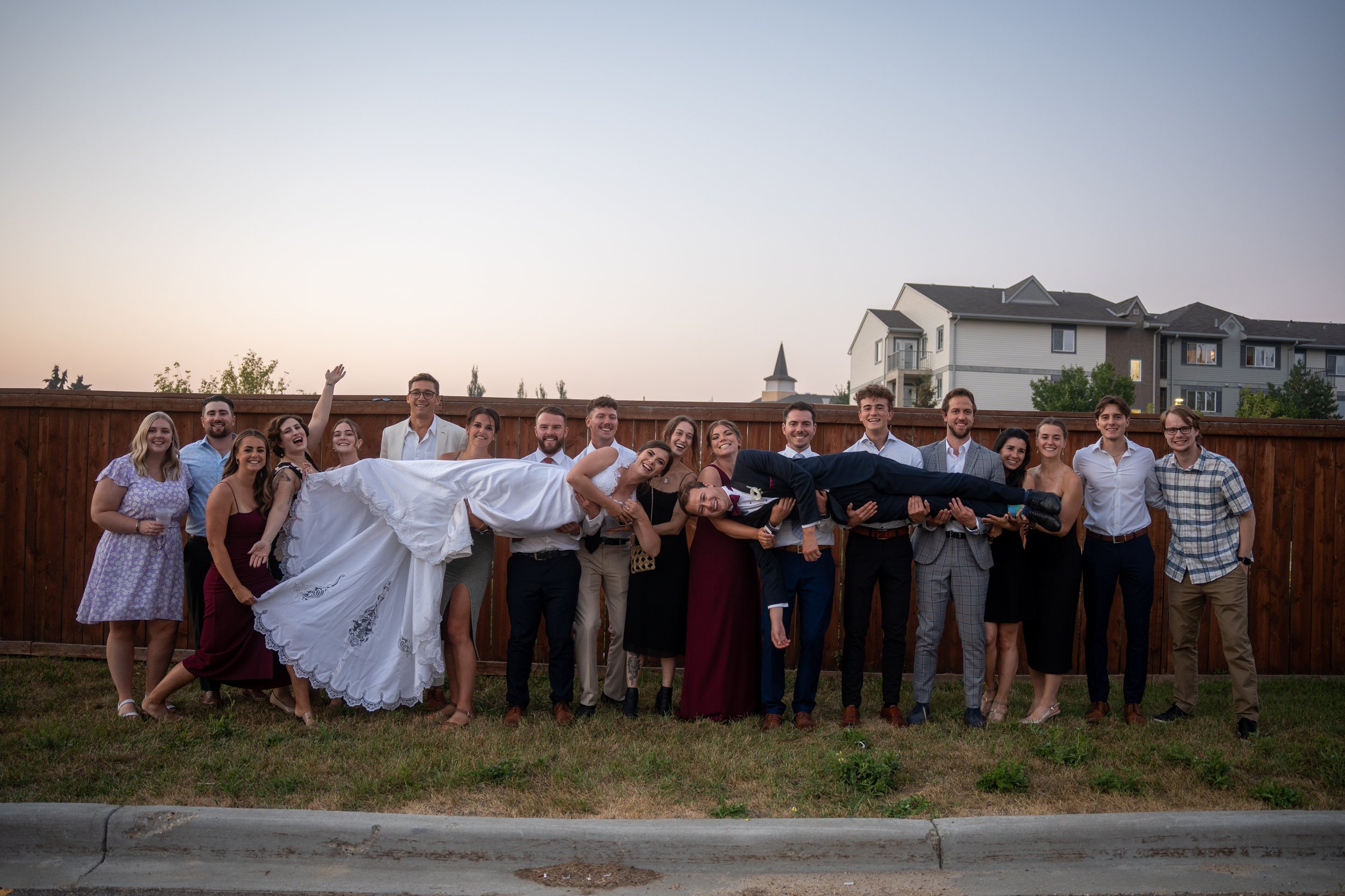 Group of people standing in front of a wooden fence with a house and a church steeple in the background, during sunset. The group is celebrating, with some smiling and one person waving.